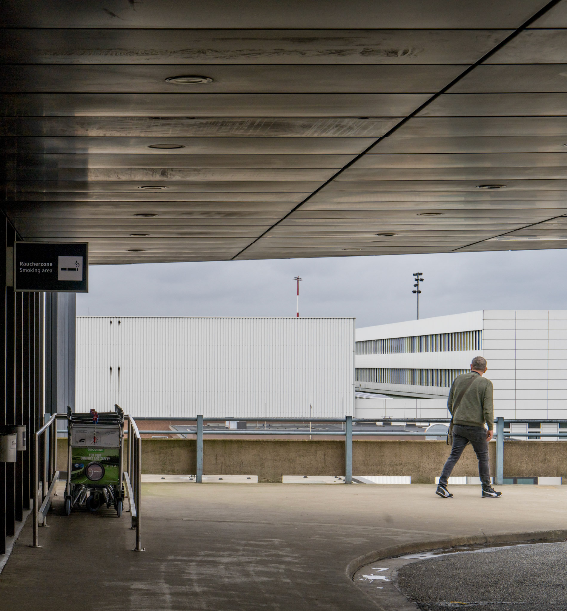 beneath the clinical gaze of fluorescent lights and the muted chatter of distant travelers, a lone figure traverses the urban expanse of hannover airport. each step echoes a subdued defiance against the rigidity of his surroundings. this is a dance of light and shadow, a fleeting moment captured where the vast, industrial structures meet the quiet solitude of a passerby. the dark overcast sky seems to weigh down, yet he moves unburdened, towards a horizon obscured by man-made giants. the rain has just left its gloss on the tarmac, reflecting not just the man but also the mood of this scene: introspective, a bit melancholic, yet undeniably hopeful.