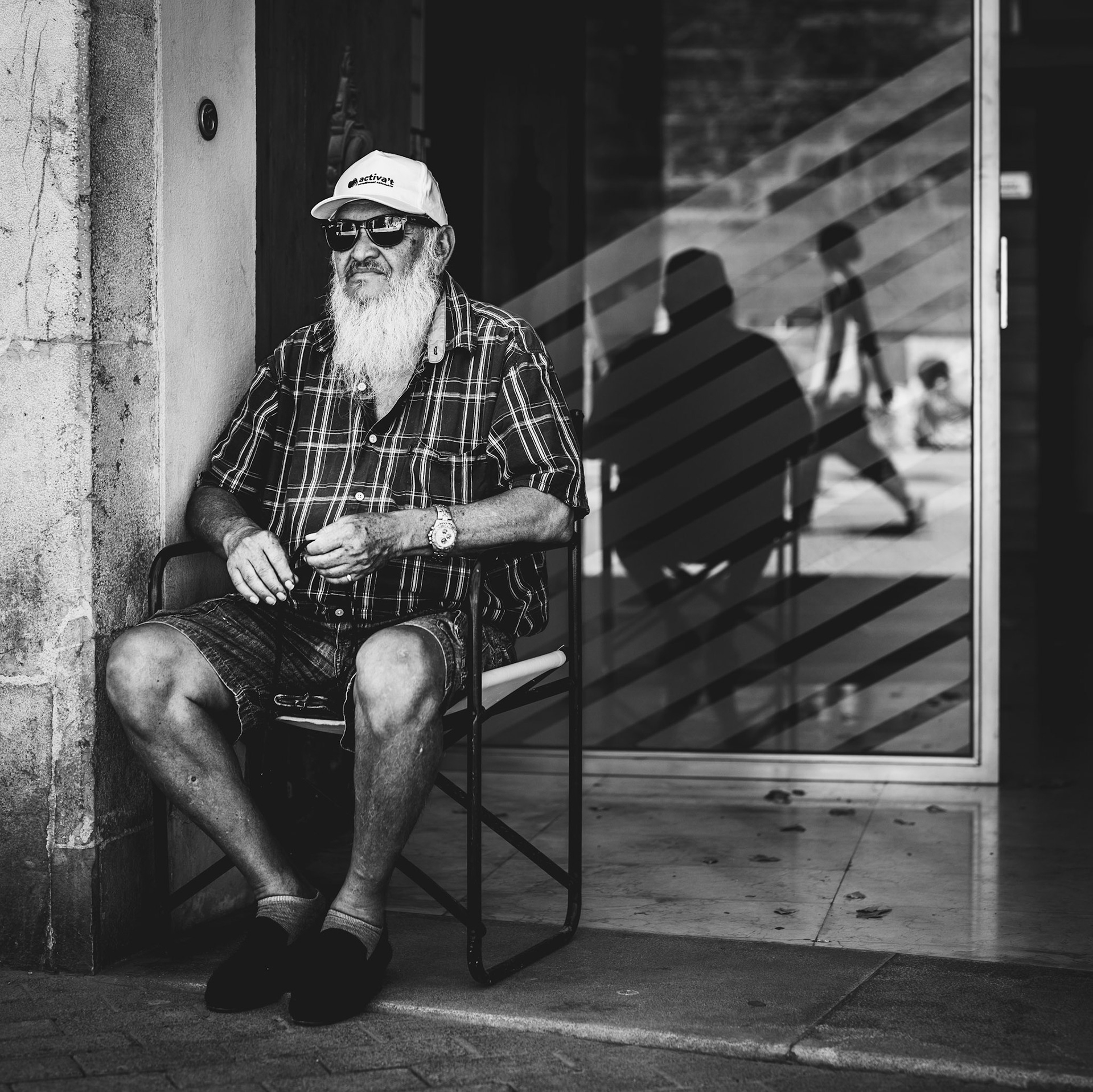 the man sits still, beads moving slowly through his fingers, each one marking a thought or a prayer. his quiet reflection contrasts with the world behind him, reflected in the glass as people move in and out of the frame. he isn't just lost in thought—he's engaging in a spiritual act, using a prayer chain to ground himself in the moment. the stillness of his body and the movement of the beads create a sense of peace amid the heat and light of the day. this was captured at plaça de la mare de déu de la salut in palma, a quiet spot where the noise of the city seems to soften, and his inner world takes center stage. time seems to slow down, as he connects with something deeper amid the city's quiet hum.