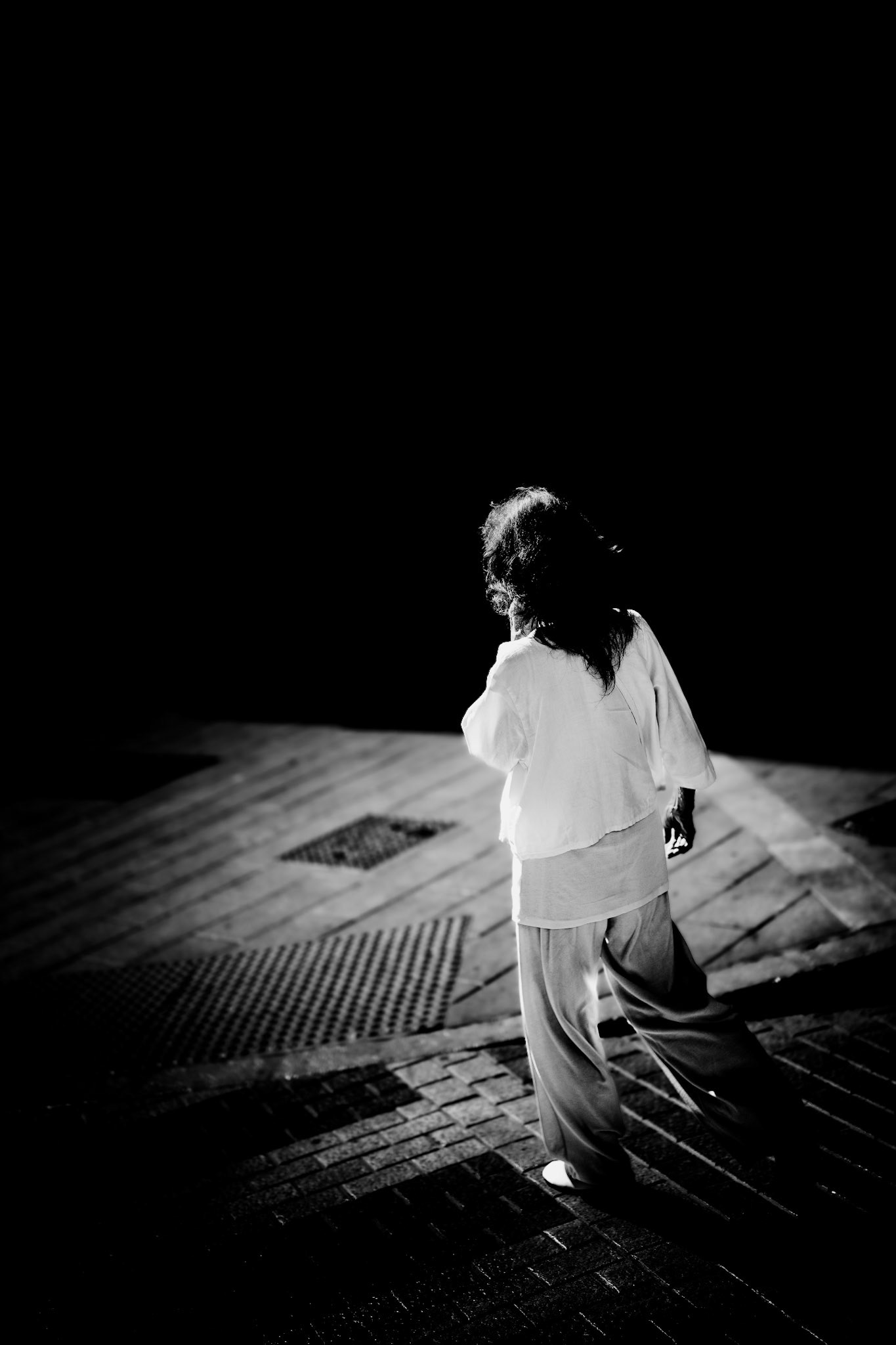 bathed in the stark contrasts of morning light and deep shadows, a lone woman strides purposefully through the streets of palma. dressed in white, her movement is swift and unyielding, undeterred by the interplay of light and darkness that envelops her. each step is decisive, driven by an inner resolve and a clear sense of direction. the avenues near plaça espanya become a mere backdrop to her determined journey, where the nuances of light and shade seem irrelevant to her focused path.