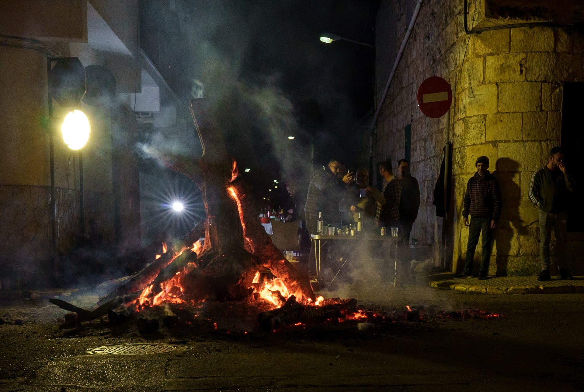 in the ancient streets of sa pobla, the night air thrums with the energy of san antoni's festivities. flames leap in the makeshift street fires, around which the villagers gather, a communal embrace against the dark. it's a time of barbecue and camaraderie, where the famed dimonis dance wildly, their figures flickering like shadows. as the village succumbs to a mystical enchantment, the primal beats of drums mix with the crackle of fires, weaving a spell over the huddled forms around the flames, a living tapestry of tradition and warmth in the heart of winter.