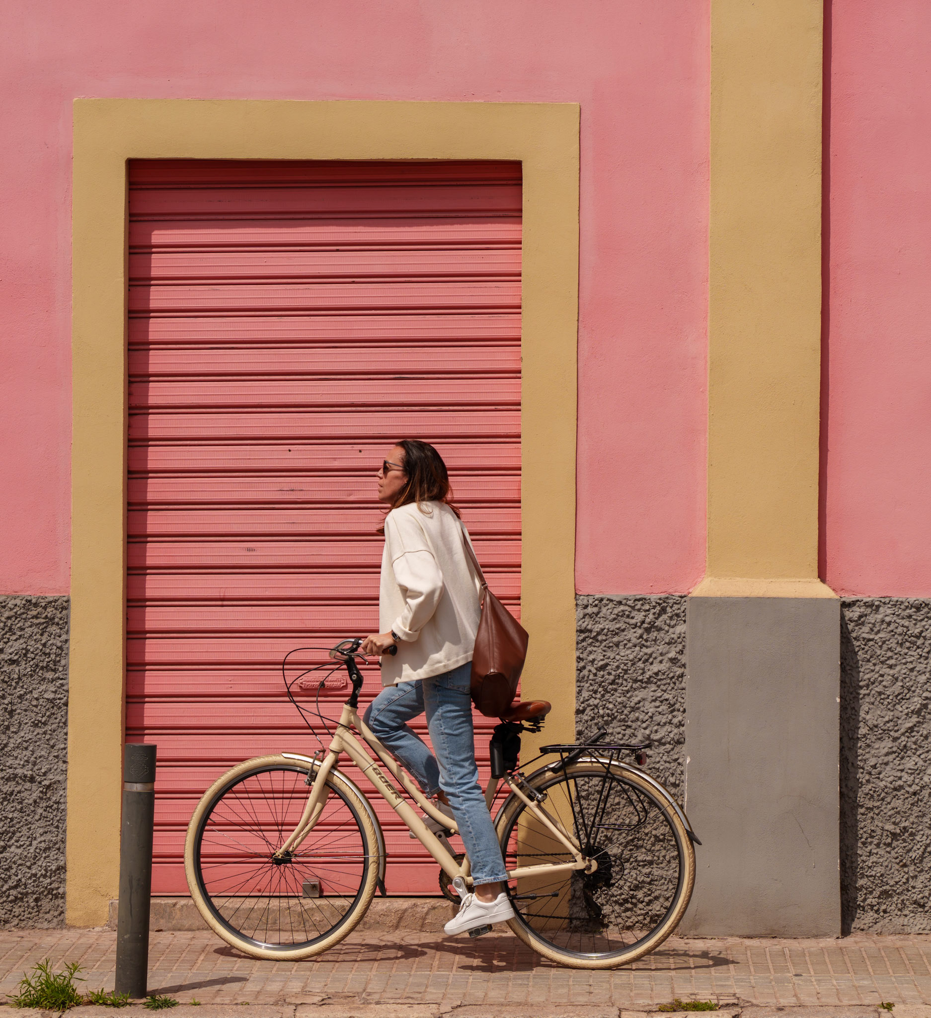 in the vibrant district of el terreno, where the quaint charm of colorful yet untouched houses whispers stories of old, she lingers. here, against a backdrop of candyfloss walls, a moment rests, suspended in the warmth of the sun. the peach blush of the rolling shutter, the mellow yellow framing it, and the coral hue of the facade, they come together like a painter's deliberate stroke on a canvas craving life. the bicycle, a pastel echo to the scene, bears a rider dressed in the softest of whites, her silhouette a serene contrast to the geometry around her. she's poised as if caught between destinations, a still life within the city's gentle heartbeat.