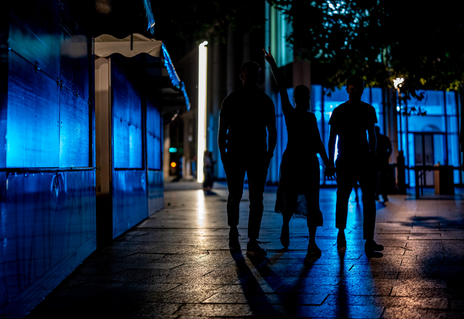 photo shows german fans returning from a public viewing after the germany vs switzerland match (1:1). silhouetted against the blue night lights, the group walks with a sense of joy and camaraderie. one fan raises a peace sign, adding a touch of celebration to the scene. the urban backdrop and the vibrant lighting create a dynamic and lively atmosphere, reflecting the spirited mood of the night.