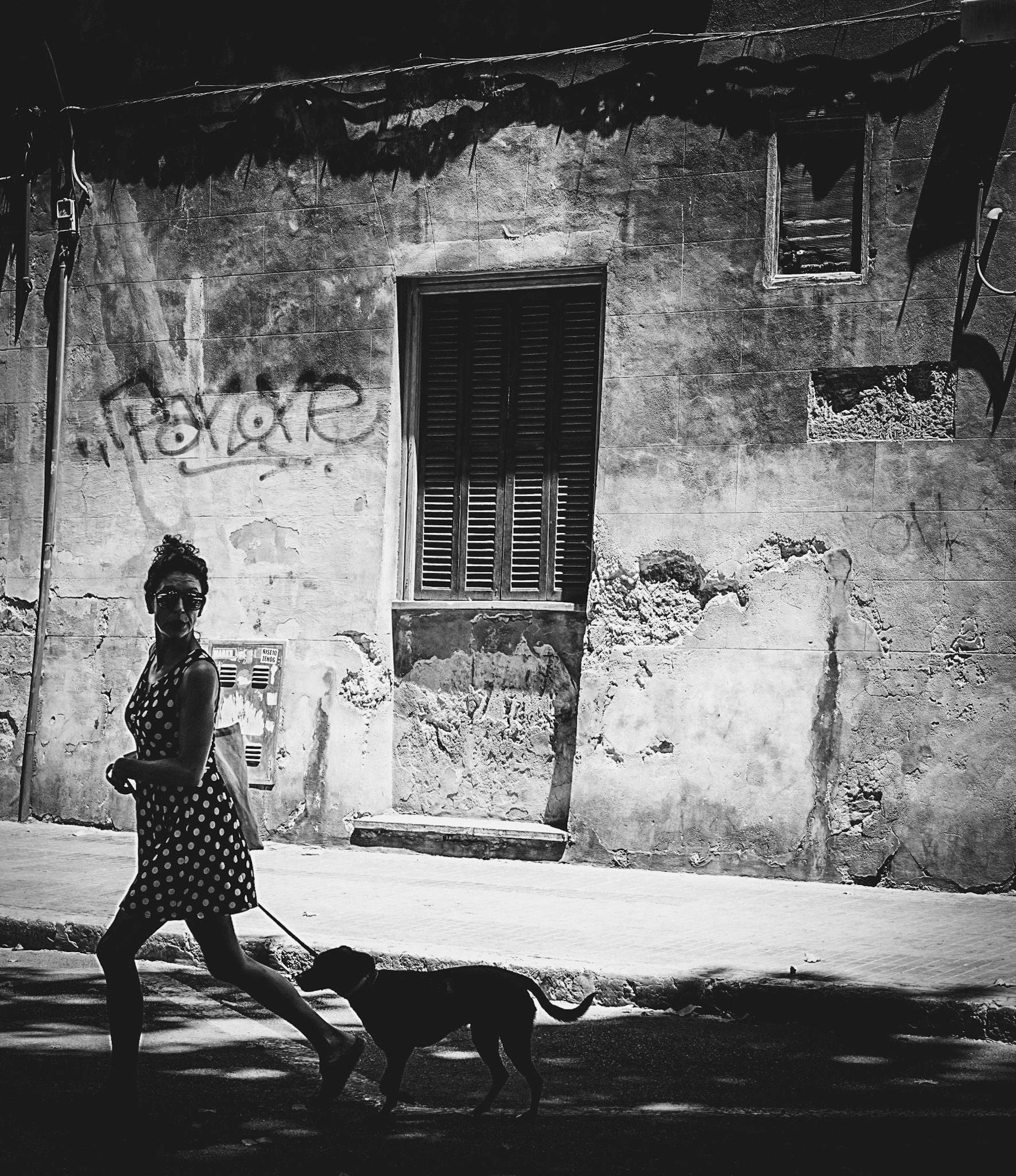 a timeless scene on the ramblas of palma de mallorca, where a woman in a classic polka dot dress strides with her dog by her side. the midday sun casts dramatic shadows against the weathered facade, creating a striking contrast between light and dark. this photograph captures the essence of street style and urban life, blending elegance and everyday simplicity in a single frame.