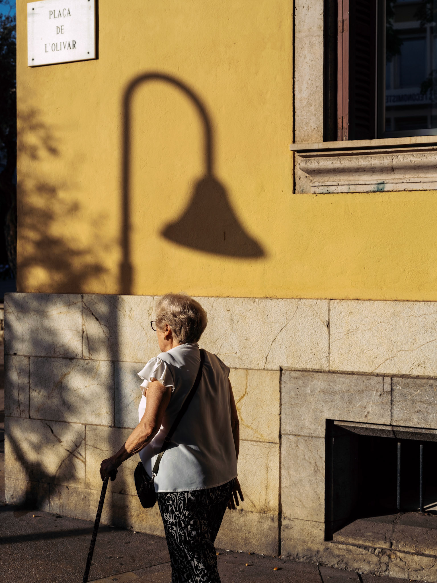 under the golden hues of the evening sun at plaça de l'olivar in palma, an elderly woman walks by with grace and purpose. her silhouette, cast sharply against the yellow facade, interacts with the elongated shadow of a street lamp, creating a visual narrative of time and age. the scene captures the essence of everyday life, where each shadow tells a story of the passage of time and the beauty found in daily routines. this moment, filled with warmth and quiet reflection, highlights the intersection of human presence and architectural elegance.