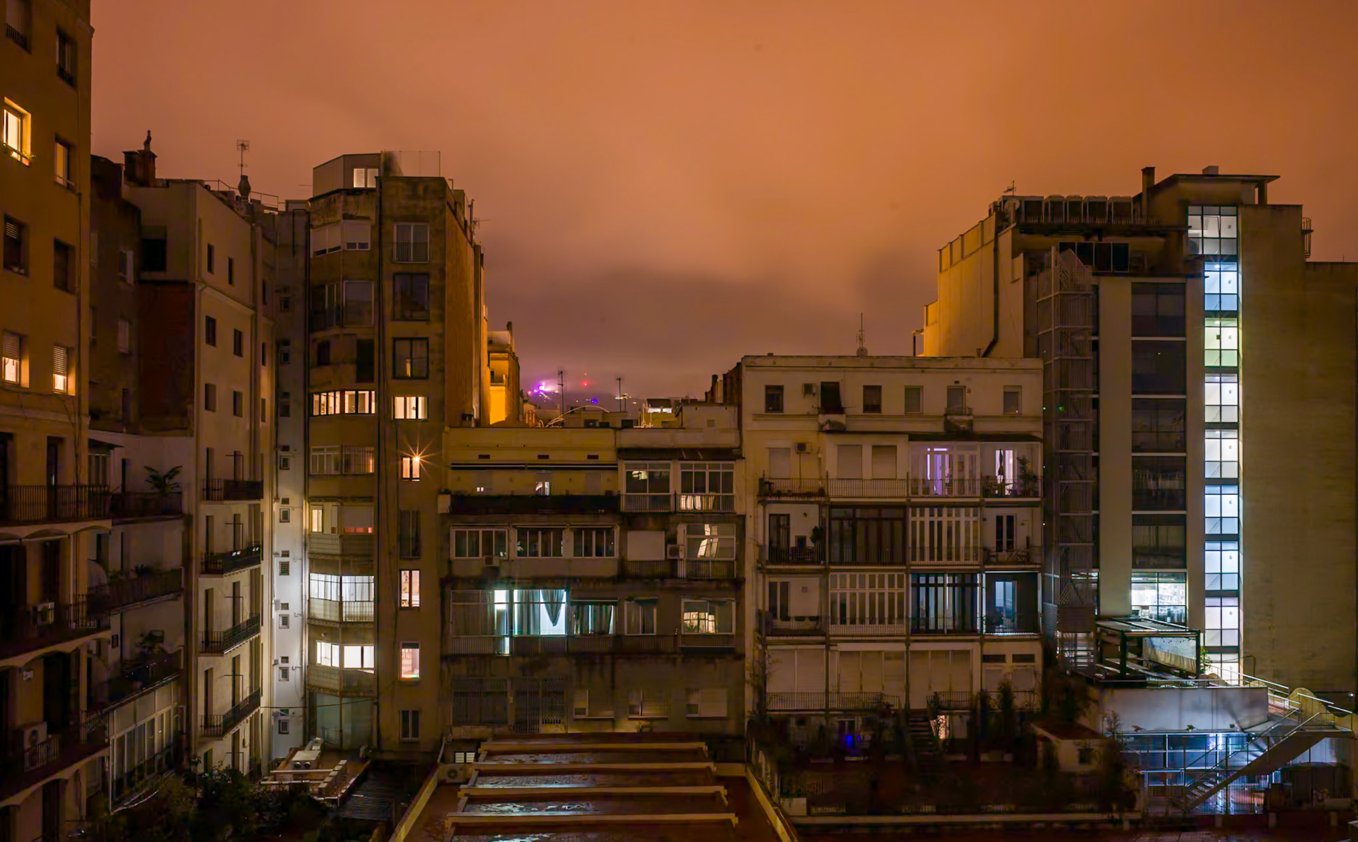 gazing out to the Barcelona nocturne, the view unfolds into an intimate tapestry of urban life. the night's canvas is splashed with warm glows from windows, each telling a different story—a mosaic of lives intersecting without meeting. the lights, like staccato notes on a dark score, punctuate the city's rhythm, the silence between them as telling as the illumination itself. in the distance, the city's pulse is a dim, purplish haze, a beacon of the nightlife that throbs unseen. in this nocturnal backyard of quiet existence, the overture of dreams plays softly, inviting the curious eye to ponder the hidden stories behind each lit window.