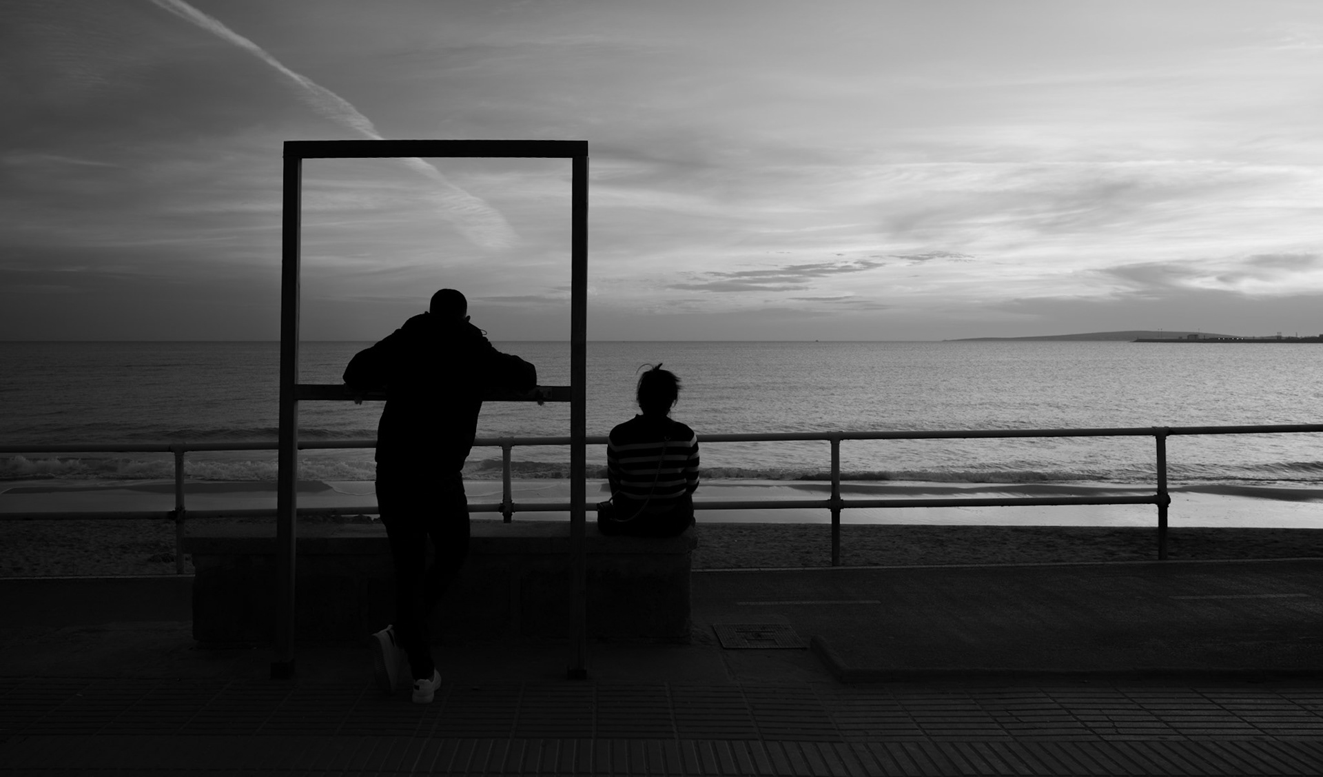 along the beach promenade of palma, silhouettes are framed not by wood or metal, but by the very horizon itself. two figures, caught in a moment of still contemplation and casual observation, share a scene where the vast sea meets the sky. it's a canvas that breathes, a living picture that evolves with the ebbing tide and the fading light. in the simplicity of this encounter, there's a narrative of shared human experience, a silent acknowledgment of life's expansive beauty, all witnessed against the grand backdrop of nature's artistry.