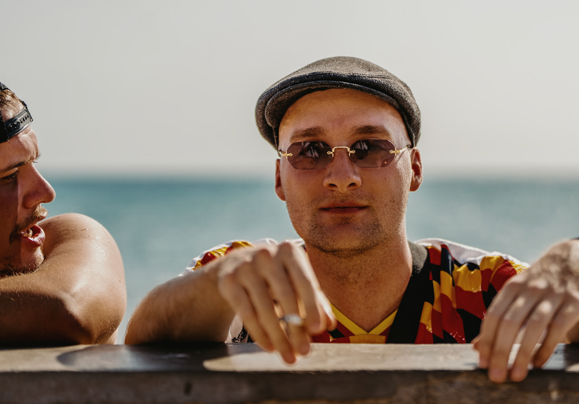 captured during a sunny day at el arenal, this candid photograph portrays two german tourists engaging in a lively conversation by the wall that separates the beach from the promenade. the casual ambiance, highlighted by the bright sunlight and the shimmering sea in the background, emphasizes the relaxed atmosphere of a seaside vacation. the expressions and body language of the subjects reflect a moment of connection and enjoyment, encapsulating the essence of summer leisure and friendly interactions.