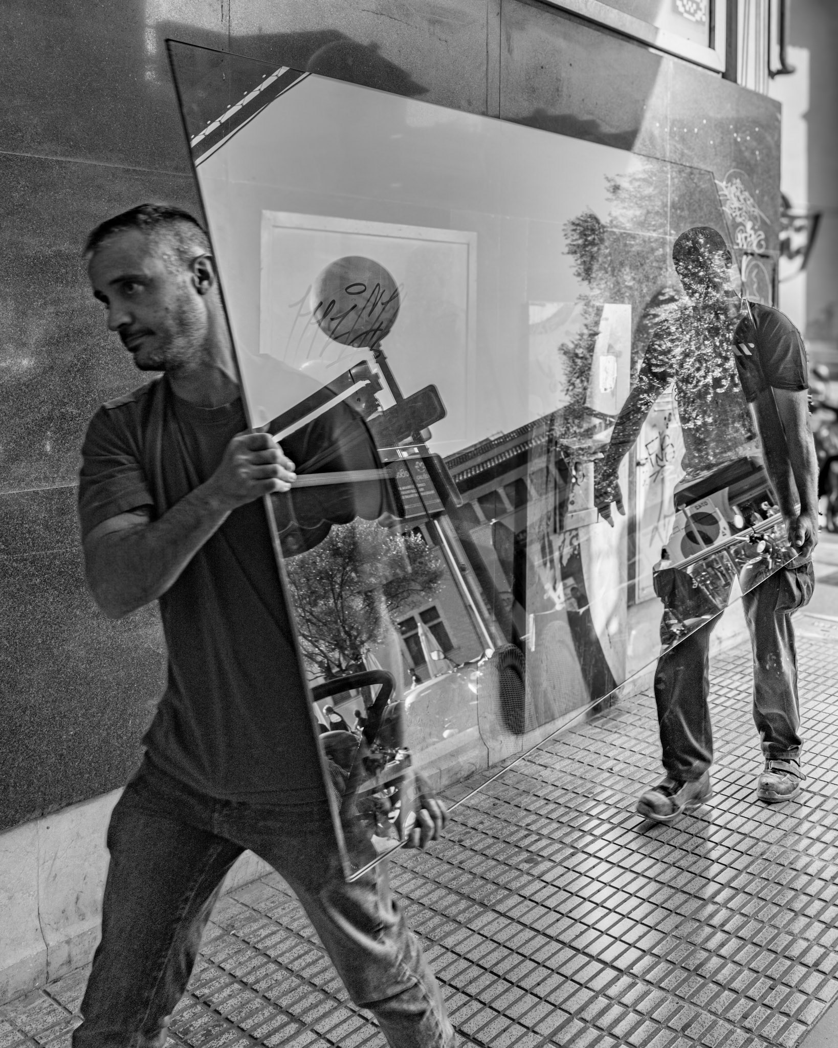 captured during a quiet moment in palma’s galeria velázquez, just a step away from the bustling mercat de l’olivar, this photo unveils a poetic intersection of reality and illusion. two workers, burdened yet graceful, carry a pane of glass that reflects not only the surrounding streetscape but also fragments of their own presence. the transparency of the glass merges their efforts with the cityscape, crafting a seamless visual dialogue between the physical and the ethereal. this fleeting moment, filled with symmetry and texture, tells a story of labor as art—where even the most mundane of tasks becomes a canvas for reflection and interpretation.