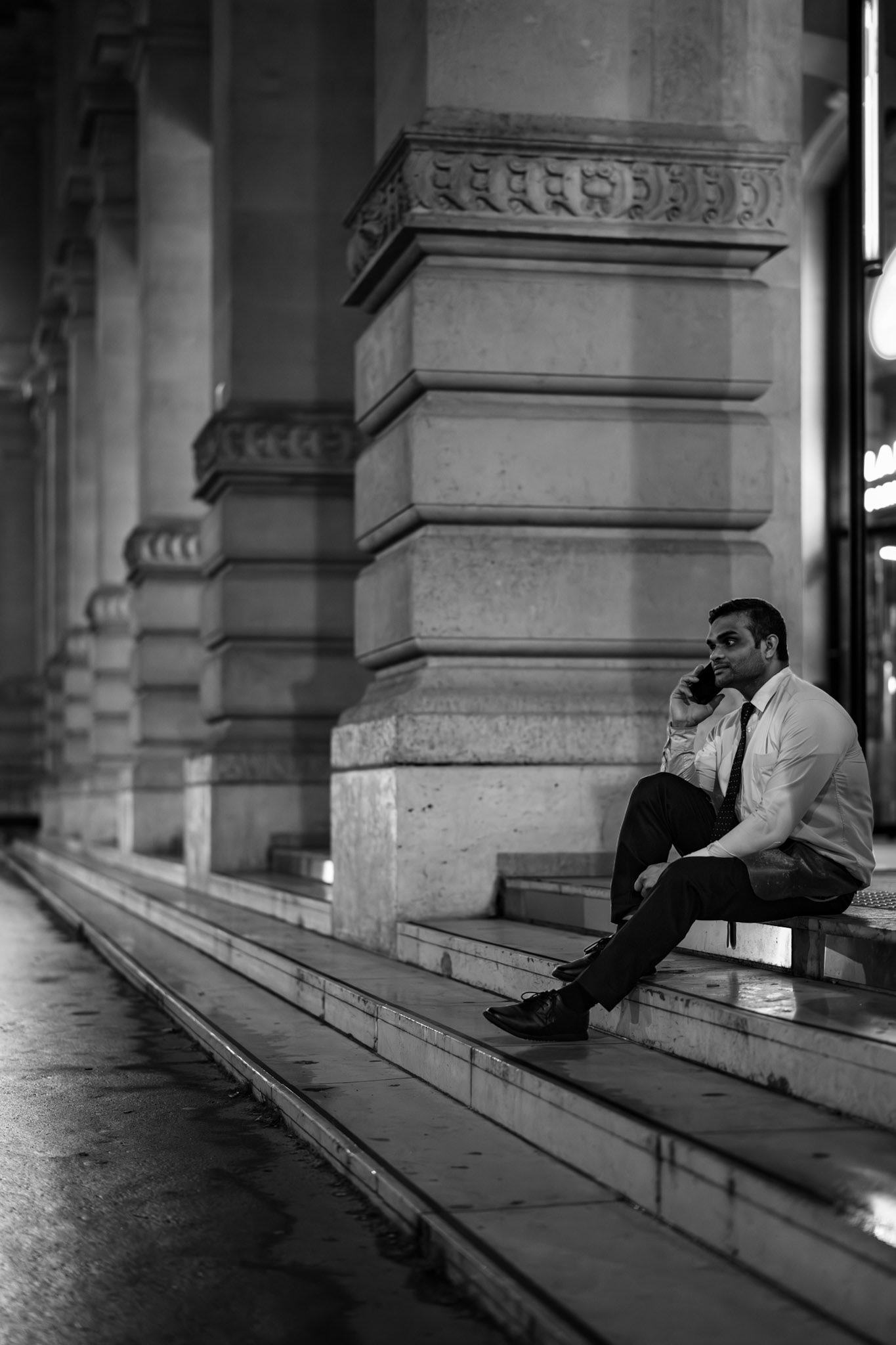 in the gentle glow of the parisian night, he sits there, absorbed, his thoughts drifting far away, tethered to another world by the phone in his hand. the grand pillars around him amplify his quiet presence, as if he's performing in a theater of solitude, a scene in black and white framed by the whisper of streetlights and the cool marble gleam of the steps.
