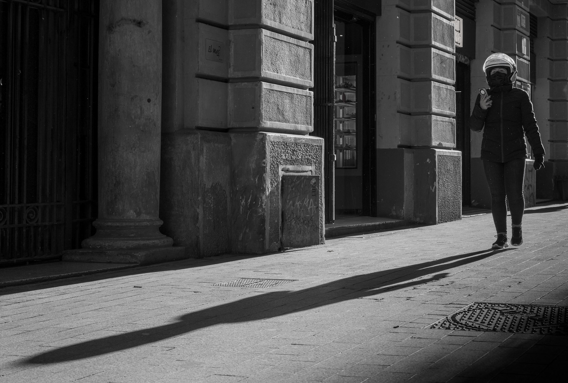 captured in the old town of barcelona, this photograph titled "landing of the aliens" plays with shadows and light to create a mysterious, almost otherworldly atmosphere. the elongated shadow of the woman in the helmet walking past the old buildings adds to the intrigue, evoking a sense of alien arrival. the use of black and white enhances the dramatic contrasts and textures, highlighting the historic architecture while adding a timeless quality to the scene. this image blends elements of street photography with a touch of surrealism, inviting viewers to ponder the story behind the shadows.