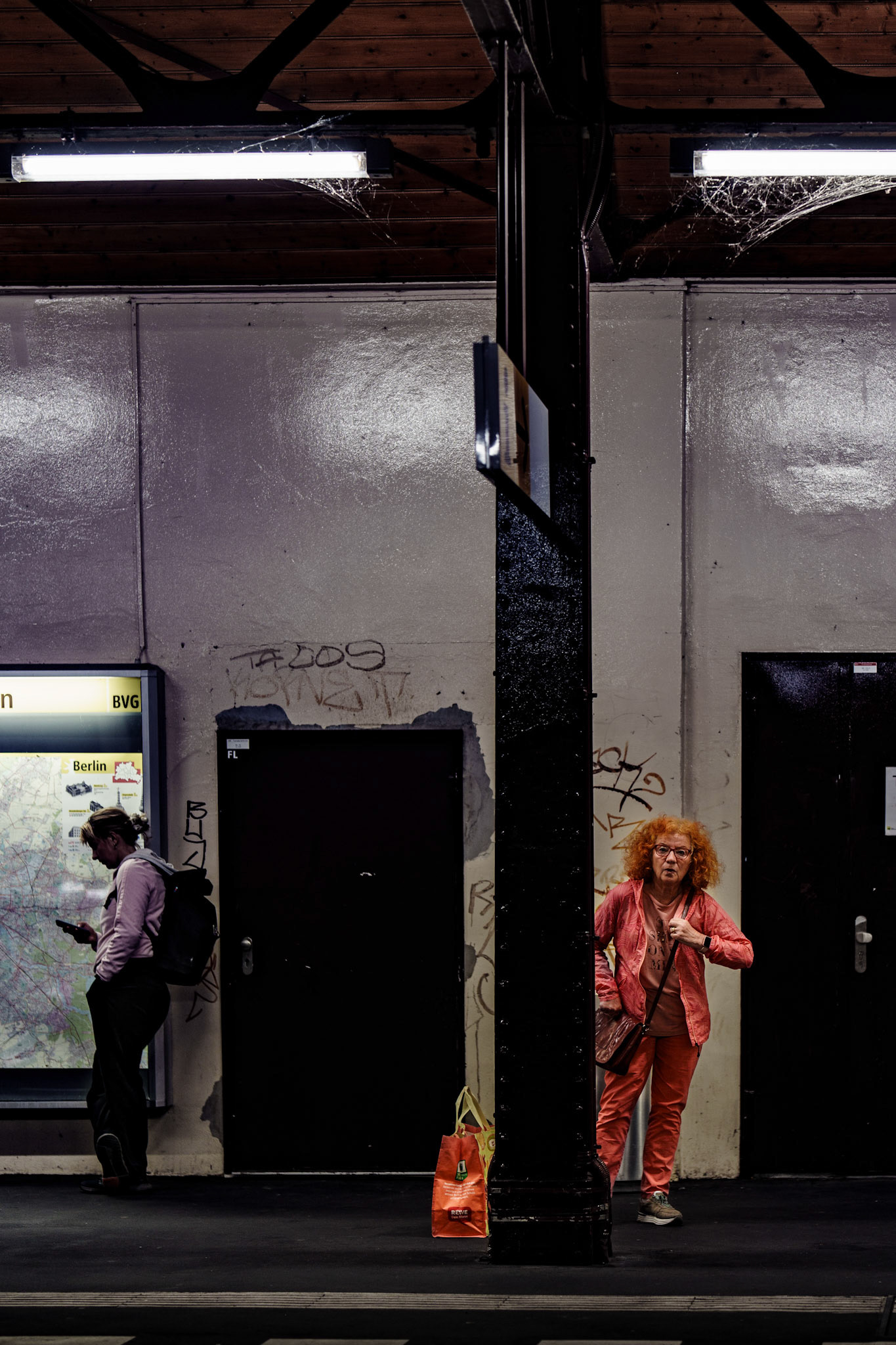 captured at schlesisches tor, berlin’s elevated station where the tracks rise above the streets, this moment blends contrasting emotions. the woman in red stands boldly against the urban backdrop of graffiti and metal pillars, her expression a mix of surprise and resilience, while a commuter to the left, absorbed in his phone, fades into the daily rhythm of the city. schlesisches tor, known for its elevated structure, casts a shadow over those below, creating a scene both mundane and extraordinary, marked by the vibrant and the unnoticed crossing paths.