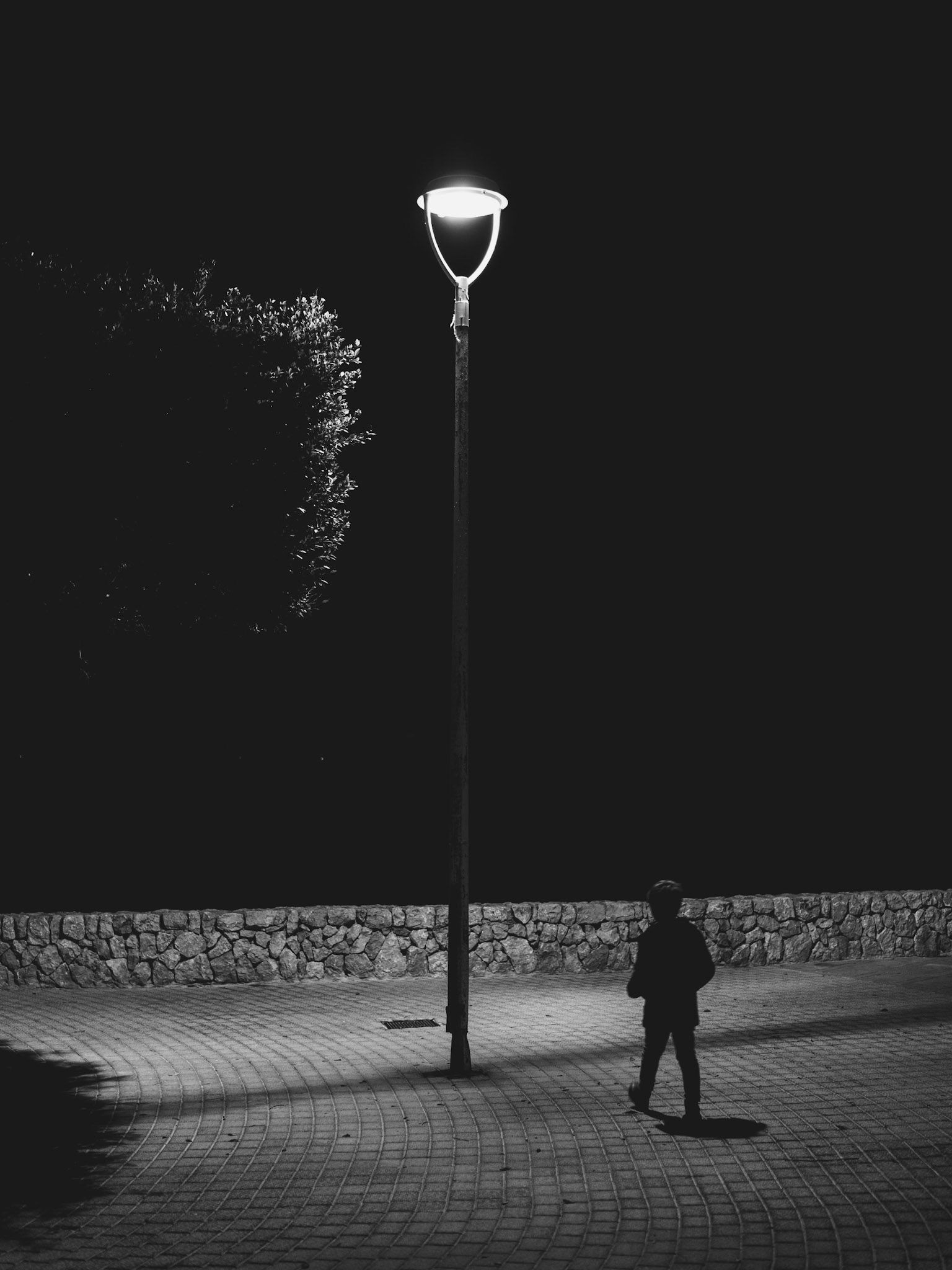 in a small park at santa ponca, a boy stands under a streetlight's glow. the darkness of the mediterranean night envelops everything but the lamp's bright beam, casting a long shadow on the brick pathway. it's a moment of quiet reflection by the sea, where silence speaks volumes.