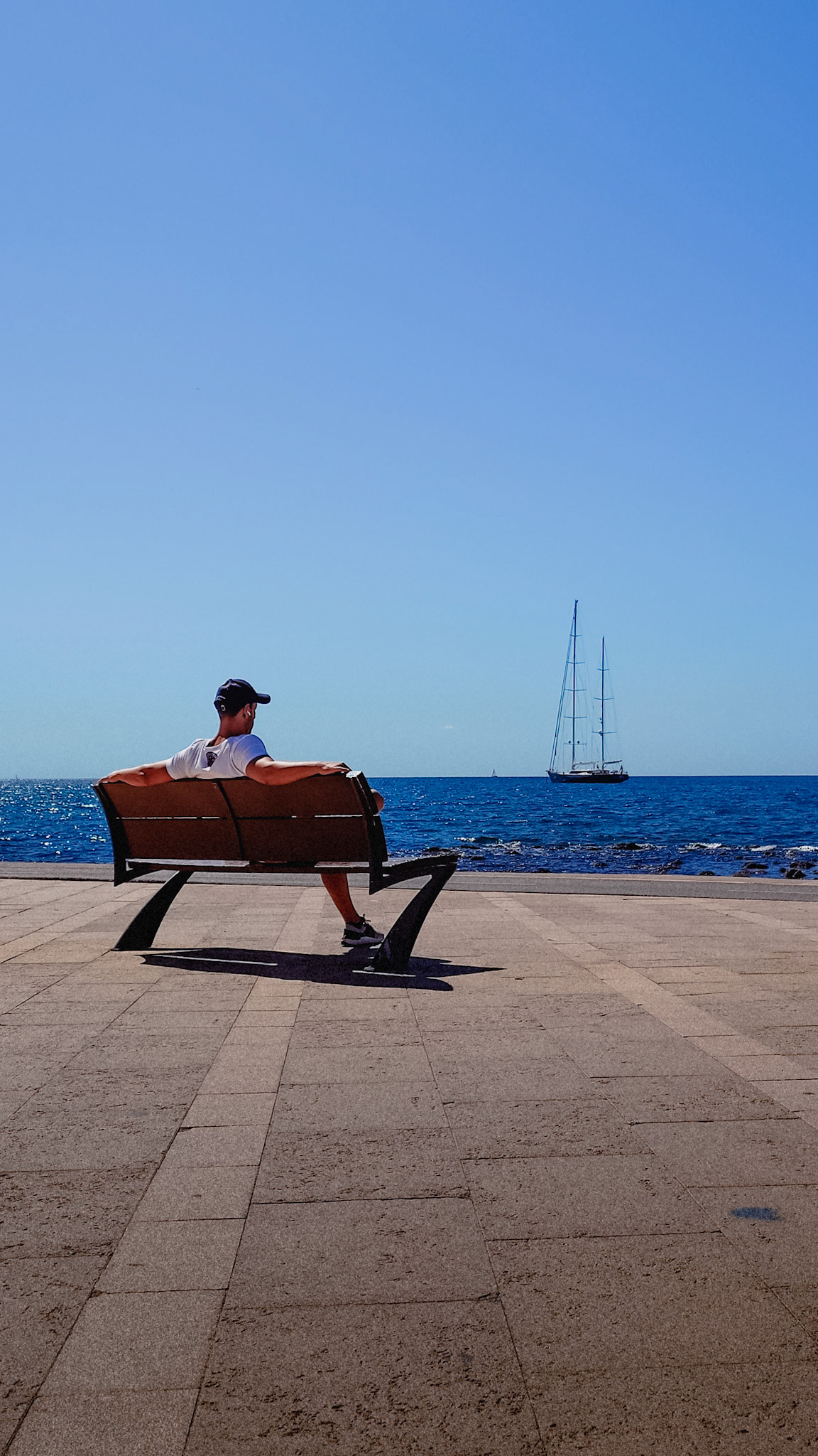 a man sits leisurely on a bench by the sea, gazing out at a sailboat gliding across the sparkling blue waters. the clear sky extends above, and the peaceful seaside setting invites a moment of reflection. the vibrant colors of the scene emphasize the beauty of a day at the sea.