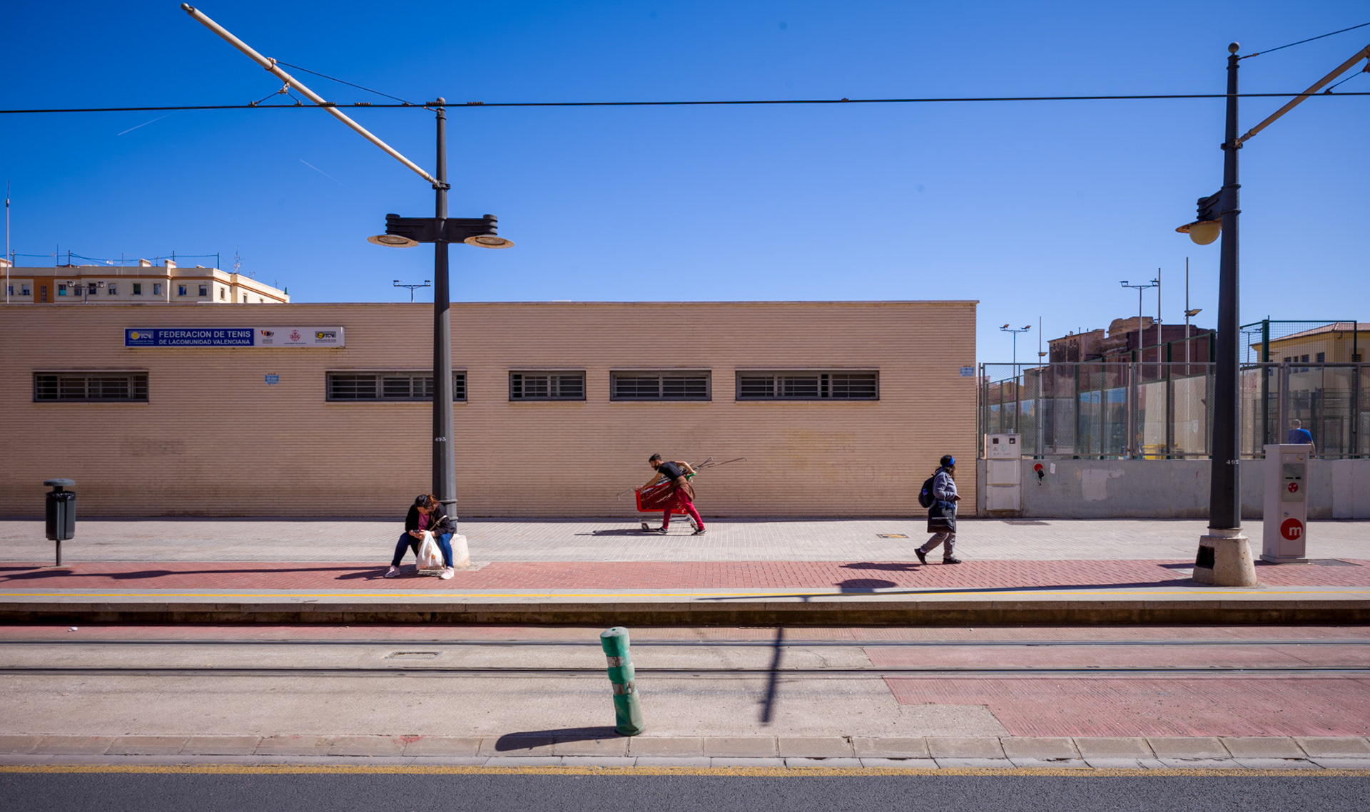 a glimpse into the ebb and flow of cabanyal, this photograph captures the essence of stillness amidst the hustle. three solitary figures, each absorbed in their own narrative, stand in contrast to the stillness of the tennis federation building behind them. in the foreground, the vibrant streaks of red and blue attire add a dash of color to the earthy tones of the urban landscape. tram lines above, like silent observers, sketch the vastness of the valencian sky, hinting at the rhythm of a city in constant motion. it’s a candid frame, where life pauses momentarily allowing us to ponder the quiet stories that unfold on the sunbathed pavements of this vibrant district.