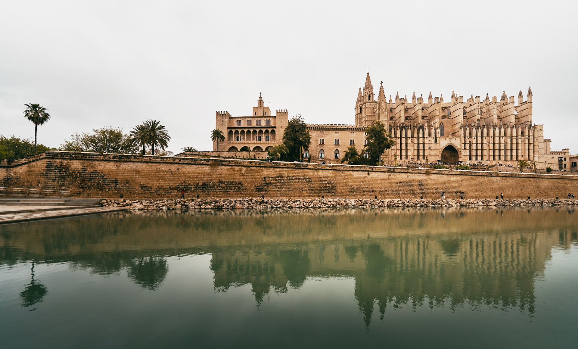 overcast skies whisper to the stoic grandeur of palma's cathedral, its gothic contours sketched against the canvas of time. raindrops tap dance on the parc del mar, crafting a symphony of ripples that mirror the edifice's ancient walls. the cathedral, a sentinel of stories, stands as an echo of devotion, where each stone is a verse of faith meticulously carved by hands long gone. here, the past converges with the present, witnessed by the casual stride of locals and the awestruck gaze of wanderers. in this tableau, the water is not merely a surface but a lens, casting the reflection of a bygone era into the gaze of today, as the elements adorn the scene with a patina of reflective tranquility.