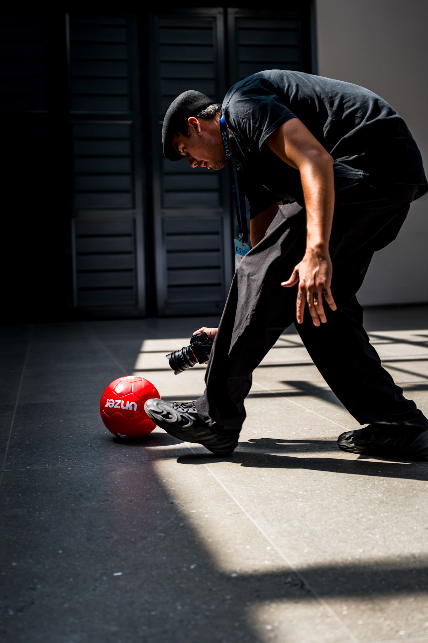 captured at the k5 conference in berlin, this photo showcases a videographer using a red ball and an ultra-wide-angle lens to achieve the perfect shot. The interplay of light and shadow emphasizes the focus and professionalism of the videographer, bent low to get just the right angle. The vibrant contrast between the red ball and the more subdued tones of the surroundings enhances the dynamic and creative energy of the moment.