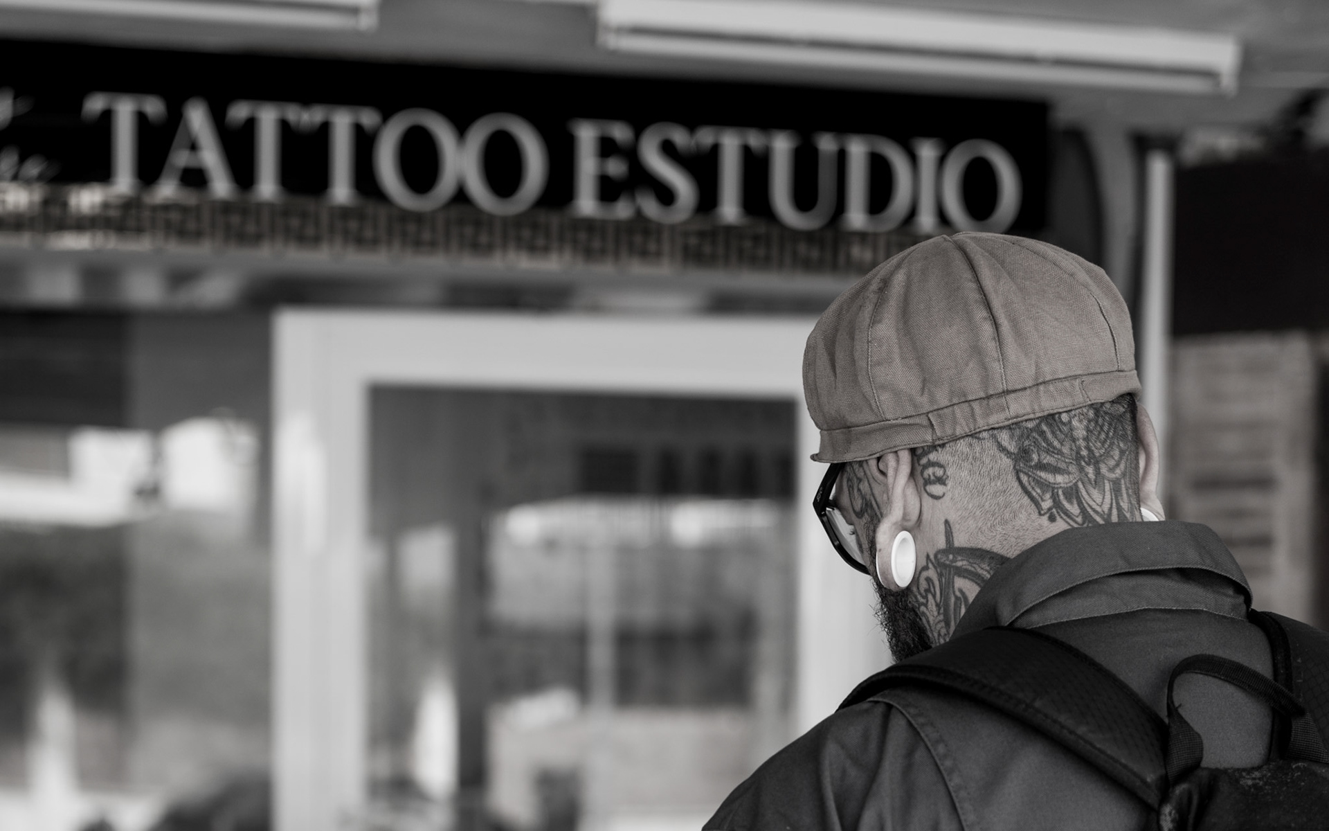 outside a tattoo studio in magaluf, a man pauses, his back to the world. the story of his journey is etched in ink on his skin, a personal gallery of life's twists and turns. each tattoo is a whisper of where he's been and who he's become. the tattoos, like a coded language, wait to be deciphered, promising tales of rebellion, loss, love, and everything in between. the man's gaze is unseen, yet one feels it piercing through the studio's facade, pondering perhaps his next piece of living art or reflecting on the permanence of the choices already made. his cap, tilted just so, adds a touch of mystery, an unspoken reluctance to reveal too much. this moment is a silent introspection, a black and white freeze-frame where stories are inked not just on flesh, but in the air around him, palpable and as real as the tattoos on his skin.