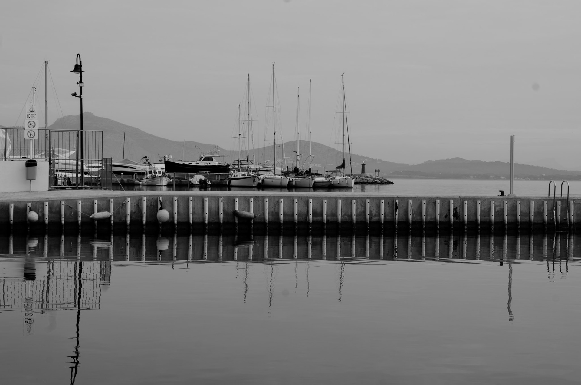 port de pollenca, draped in winter's subtle serenity, presents a scene of quiet harmony. the marina, a gallery of sails at rest, awaits the whisper of adventure. this grayscale tableau is a study of calm, where even the water seems to hold its breath, mirroring the world above in perfect symmetry. the mountains, guardians in the distance, rise softly against a sky of gentle gradients. the pier, lined with round fenders like a string of pearls, marks the boundary between the known and the beckoning sea. it’s a photograph of contrasts — the stillness of the boats against the latent power of the sea, the solidity of the pier reflected in the fluidity of the water. this moment is a silent ode to the contemplative beauty of winter by the sea, an invitation to stand still, to look closer, to listen to the hush.