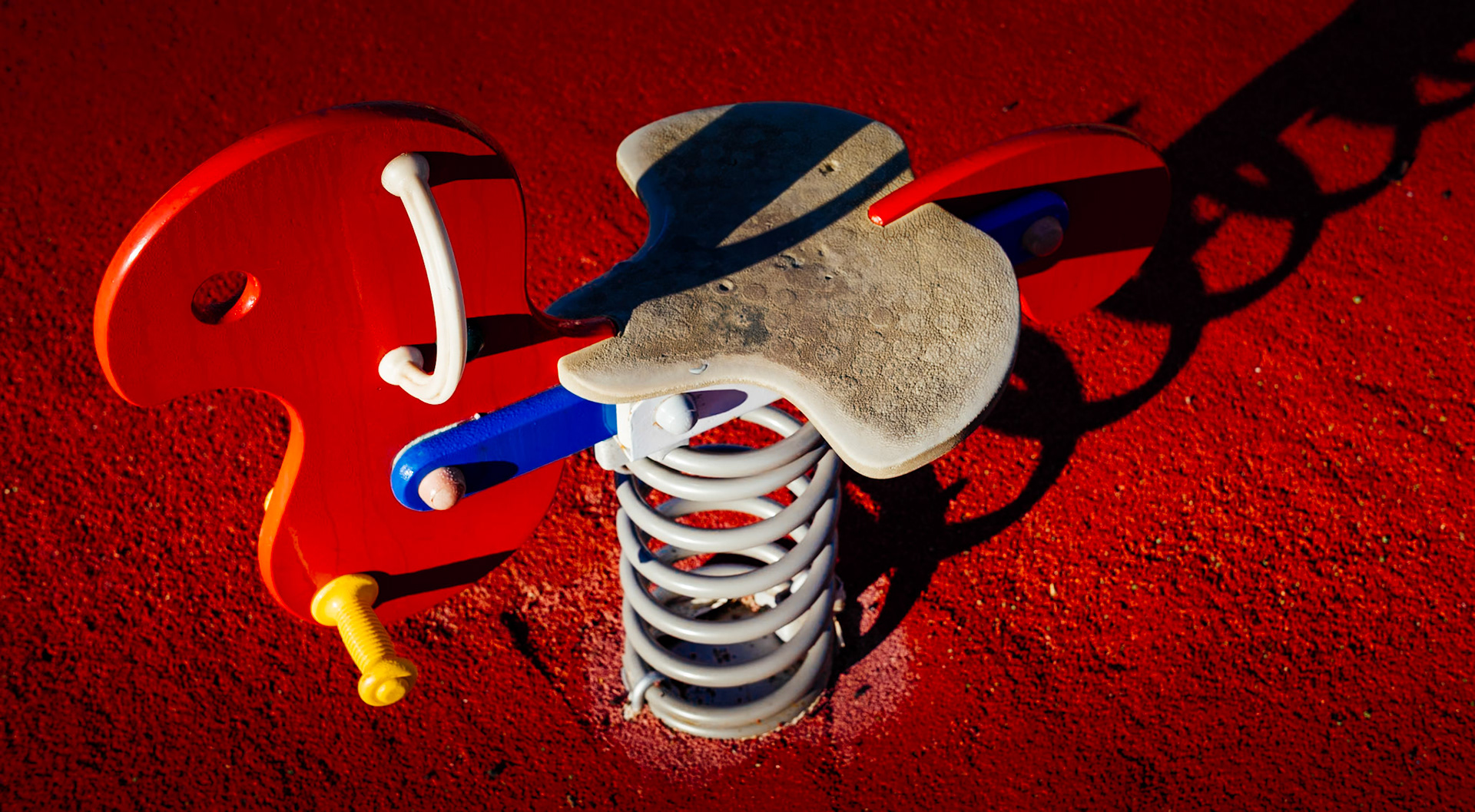 captured in palmanova, this photo is part of "a morning at the playground" series. the winter sun casts long shadows, creating a stark contrast against the vibrant red safety surface. the playground stands quiet, its colorful spring rider poised in stillness, waiting for the laughter and chaos of play that comes with the warmer days.