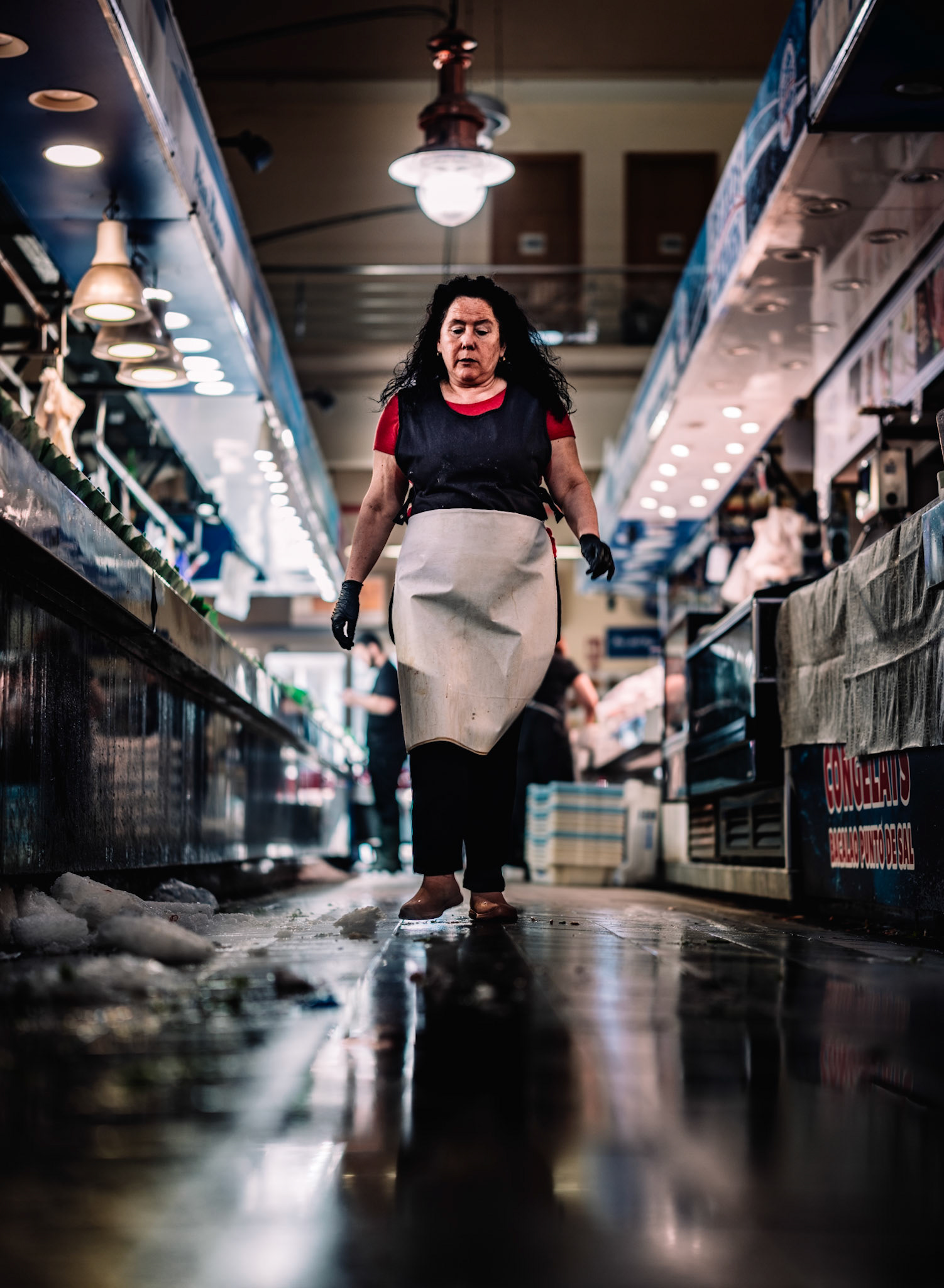 at the end of a busy saturday in the mercat de l’olivar in palma, this photo captures the moment of calm after a long workday. the vendor, in her apron and work gloves, walks slowly through the rows of market stalls, while melting ice on the floor marks the traces of the day. the warm tones of the lighting and the reflections on the wet floor create an atmosphere of exhaustion and quiet after the bustle of market activity. this image reveals the human side of the market, where hard work and dedication are behind every fish sold.