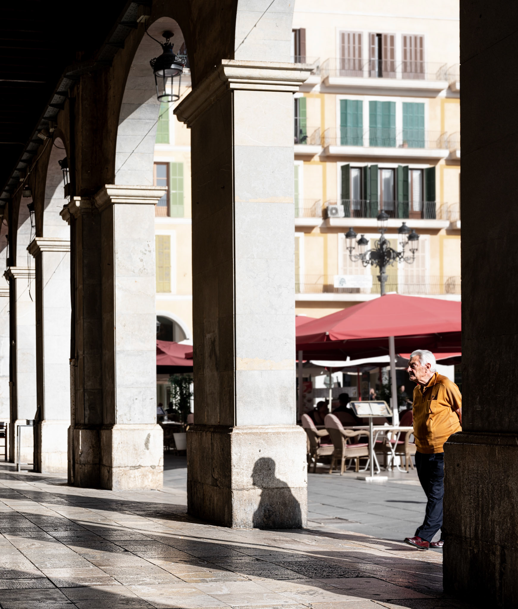 captured in palma at plaça major, this image depicts a man transitioning from the sunny plaza into the shaded arcades. his shadow is sharply cast on the stone column, adding a dynamic element to the scene. the vibrant colors of the buildings and red umbrellas in the background contrast with the cool, shaded passageway. the man's movement captures a moment of transition and contrasts with the stillness of the surroundings, highlighting the interplay of light and shadow in this lively setting.