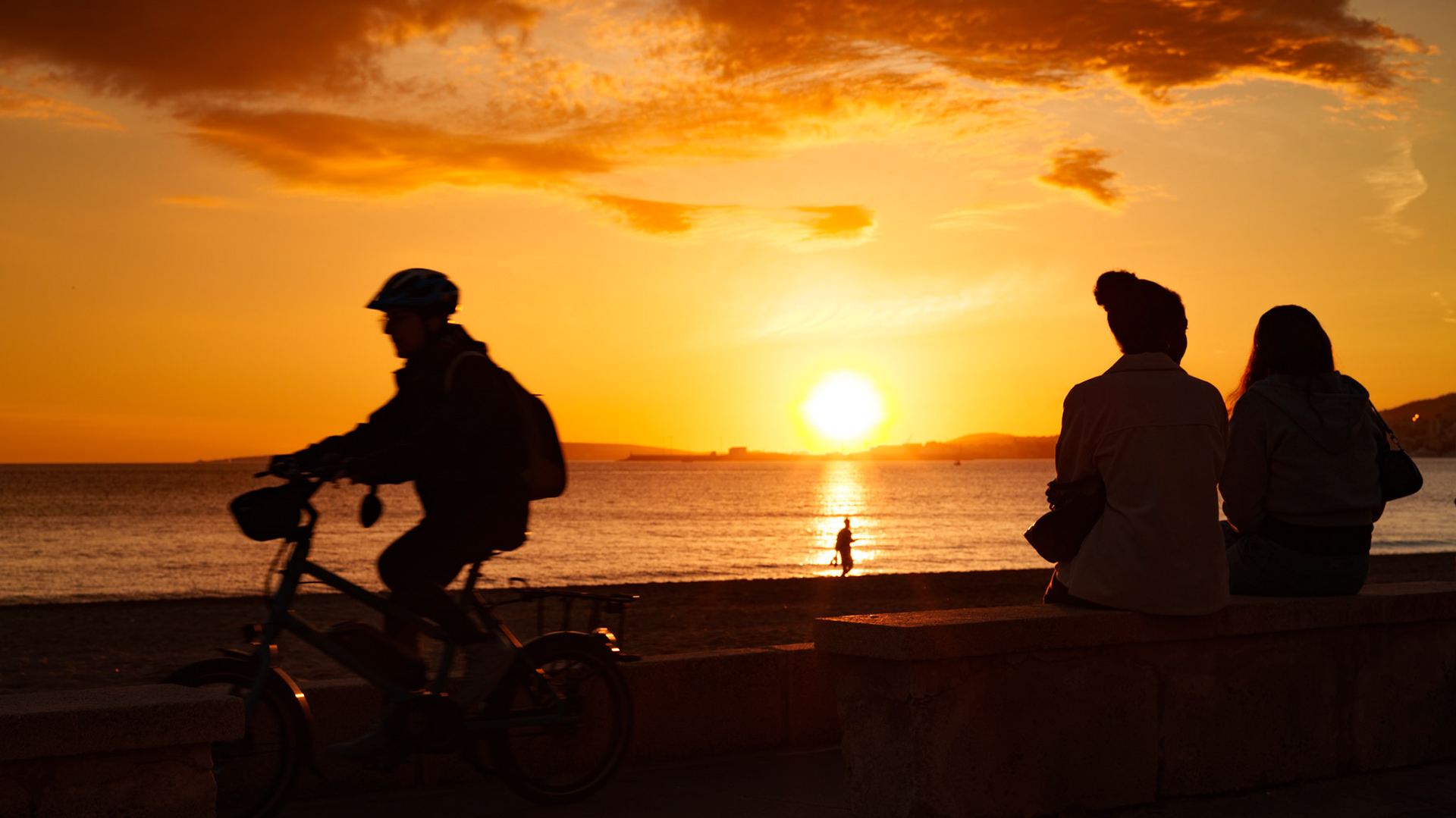 the most stunning sunset unfolds in portixol, a scenic waterfront district of palma de mallorca. silhouettes are painted against the fiery backdrop as a cyclist passes by and a couple sits, immersed in the golden hour. the sun dips low, casting a glow that turns the sea to liquid gold.
