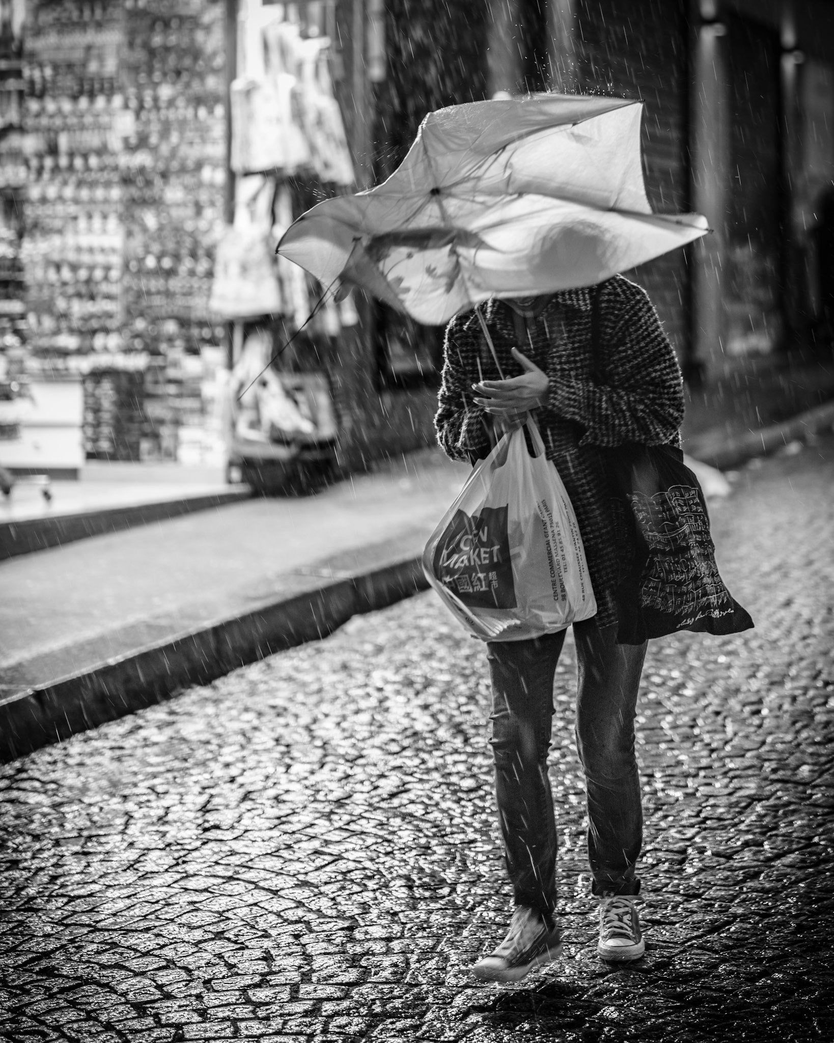 walking through the narrow streets of montmartre, the storm struck suddenly, flipping her umbrella inside out like a metaphor for the chaos of life. raindrops slashed against her like tiny daggers, bouncing off the wet cobblestones, while she held on to her shopping bags like lifelines. her face, hidden behind the unruly umbrella, became anonymous in the moment—just another figure lost in the relentless rain. the blurred lights of the city in the background seemed to mock her, offering no warmth or refuge. this photograph captures that fleeting moment when nature takes control, leaving us vulnerable and exposed, yet determined to keep moving forward.
