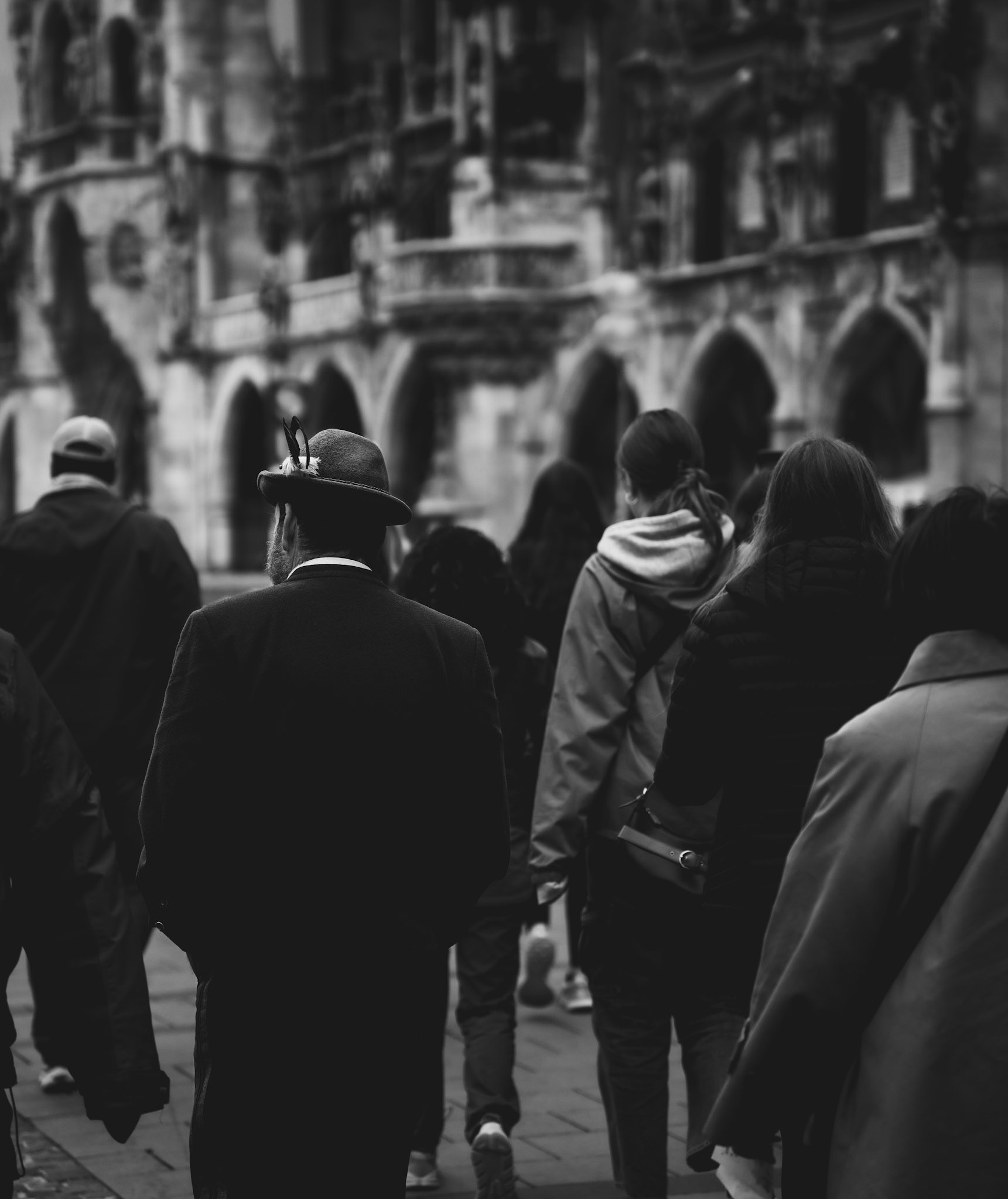 in the heart of munich's marienplatz, a lone figure in traditional bavarian attire strides through a sea of tourists. the timeless architecture of the city hall looms in the background, a silent witness to the blending of eras. the monochrome palette accentuates the stark contrast between the past and the present, the local and the visitor. this image captures a fleeting moment of cultural convergence, a scene where history and modernity walk side by side.