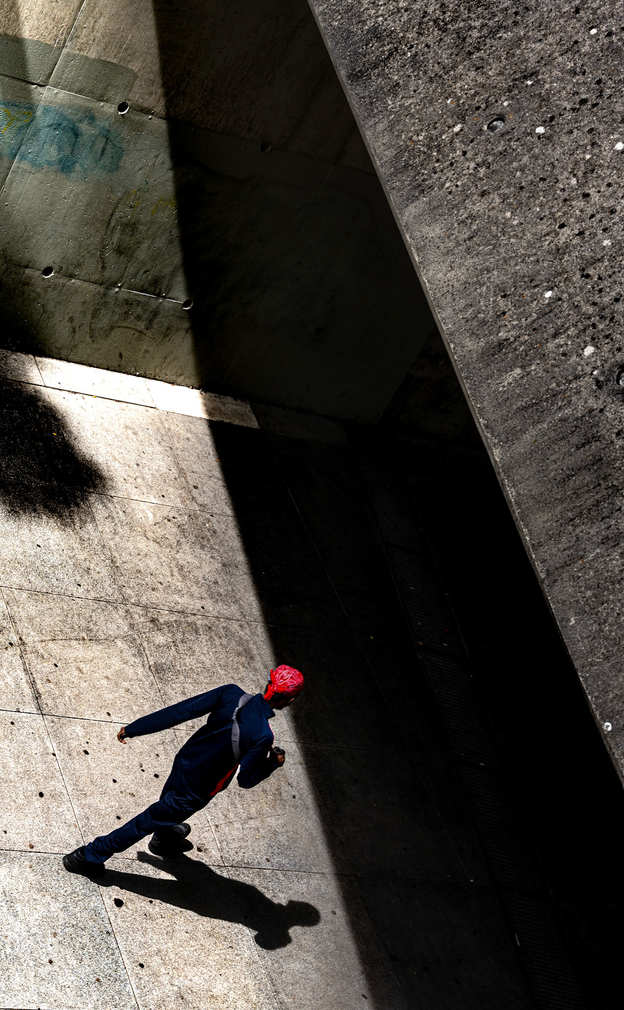 in the hidden corners of the city, where light meets shadow, a lone figure strides with purpose. the bright red of a bandana punctuates the deep contrast of concrete and darkness, a bold statement against a canvas of muted grays and blacks. from above, the perspective shifts — a solitary silhouette caught in a moment of transition, moving from the shadows into the light, or perhaps the other way around. the ground beneath, textured and weathered, tells stories of countless steps, of journeys untold. the sharp geometry of shadows cuts through the scene, creating a sense of tension and mystery. it’s a fleeting glimpse of urban solitude, a dance between movement and stillness, light and dark, captured in a moment that invites contemplation.