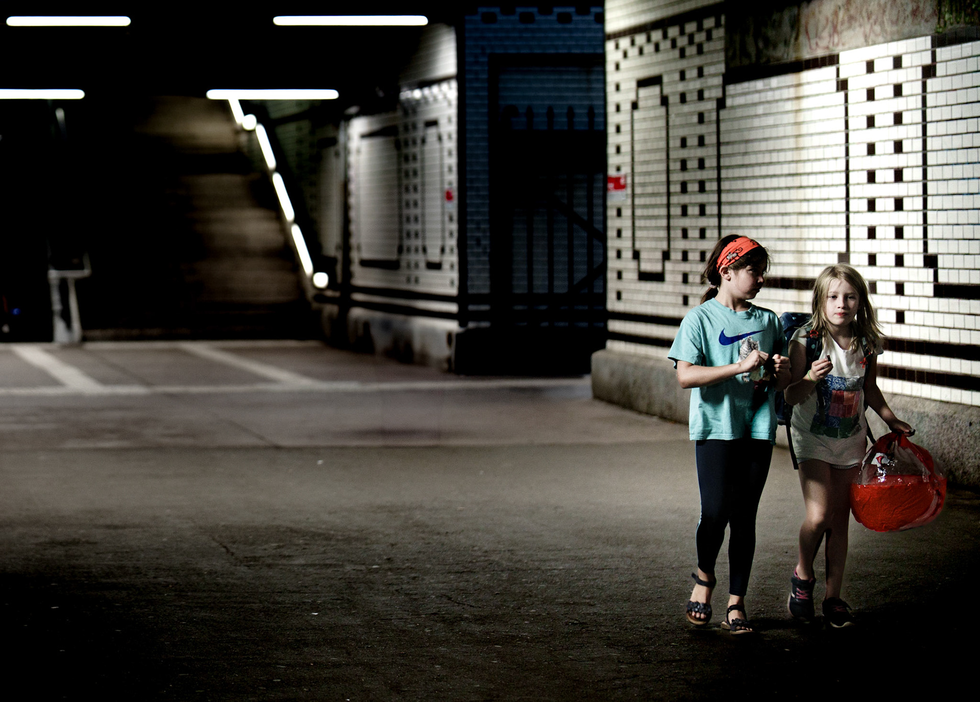 two children walk side by side, their quiet steps echoing through the dim, urban tunnel. the neon lights overhead flicker, casting faint reflections on the polished tiles. they carry remnants of the day—perhaps candy or toys—but their faces betray a mix of focus and weariness. the world around them, grimy and shadowed, feels far too vast for their small figures. they walk together, yet somehow the space between them and the city stretches infinitely. everything is still, but something about their path feels unresolved—like a passage through the unspoken loneliness of a city night.
