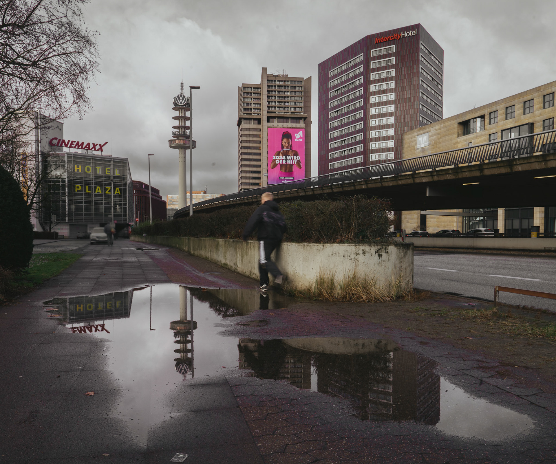 on a grey, cloudy day in february, the cityscape of hannover reveals its muted tones behind the central station. the overcast sky looms above, casting a somber mood over the urban environment. reflections in a puddle on the pavement catch the mirror image of a tower, hinting at a city in pause. a lone figure blurs into motion, juxtaposed against the stillness of the architecture and the stark outlines of the buildings.
