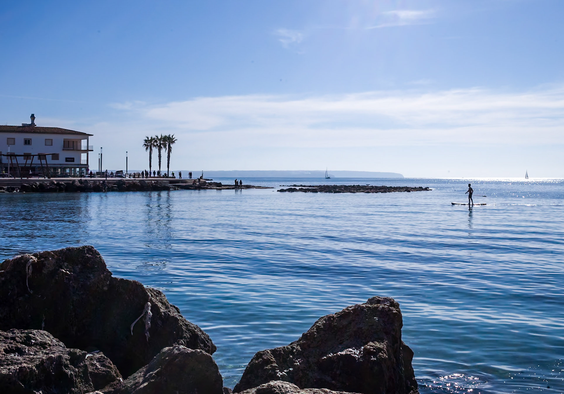 the image depicts portixol, a tranquil seaside district of palma de mallorca, bathed in sunshine on a quiet, nearly windless winter day. the serene sea mirrors the clear blue sky, punctuated by a lone paddleboarder and distant sailboats, evoking a sense of peace and leisure in this picturesque coastal setting.