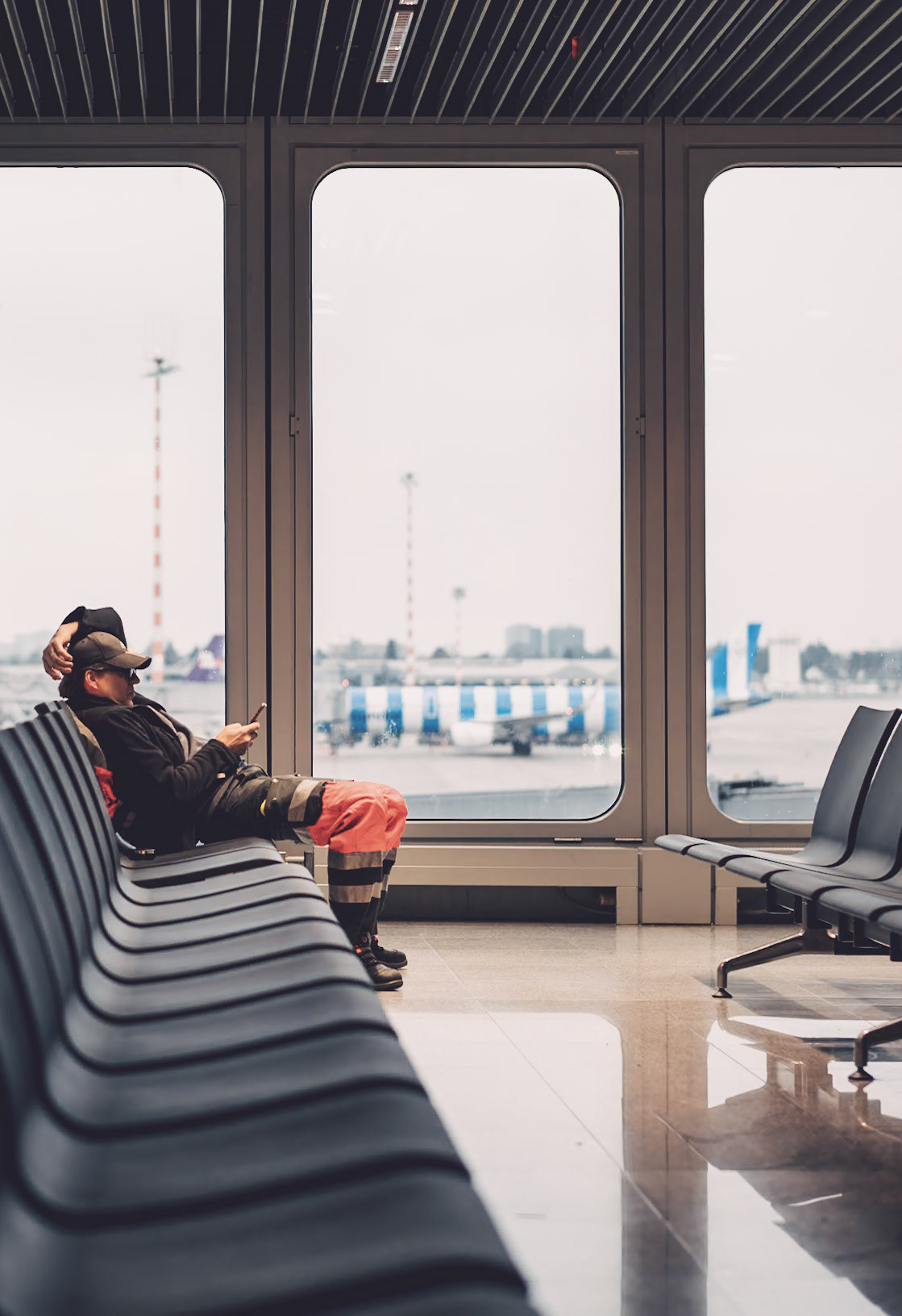 at düsseldorf airport, captivated by the interplay of lines and reflections, a man in striking orange work attire sits in contrast. the quiet terminal around him is a blend of sleek chairs and gleaming floors, a space where travelers pause, and the outside world of planes and service vehicles is glimpsed through vast windows.