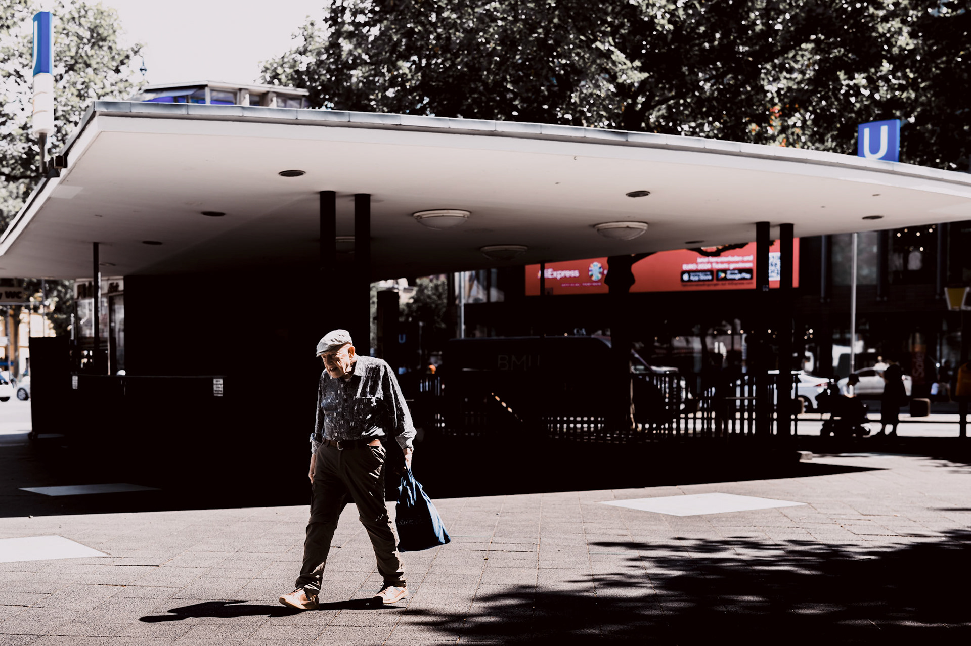 on a sunny day in berlin at kurfürstendamm, the city hums with activity. an elderly man, dressed in earth tones and carrying a blue bag, makes his way across the open space, casting a long shadow on the pavement. the entrance to the u-bahn looms behind him, its modern structure contrasting with the man's timeless presence. this image captures a moment of quiet movement in the bustling city, where every shadow tells a story and every step echoes with the history of the streets.