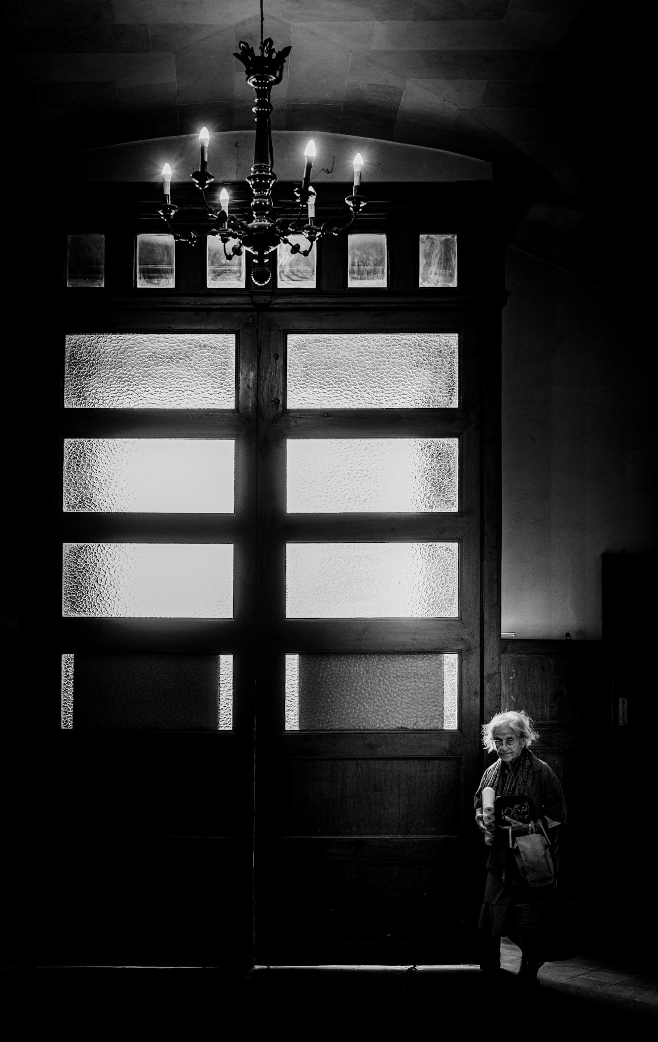 in the dimly lit vestibule of a church in palma de mallorca, an elderly woman stands, the soft glow of the chandelier casting gentle light on her weathered face. the wooden doors behind her are closed, but the frosted glass lets in just enough light to create an ethereal atmosphere. her presence adds a timeless quality to the scene, as if she holds the stories of countless years within her quiet gaze.