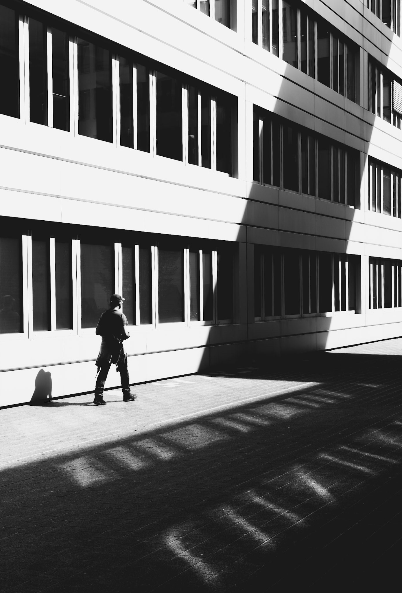 this shot captures a fleeting moment where urban geometry meets human presence, taken in a quiet corner of the city. the sharp contrasts of light and shadow paint a striking scene, where the figure is caught walking along a path defined by architecture and sunlight. the lines and shapes create a sense of rhythm and movement, emphasizing the everyday dance of city life. it’s a visual exploration of space, light, and the quiet moments that often go unnoticed.
