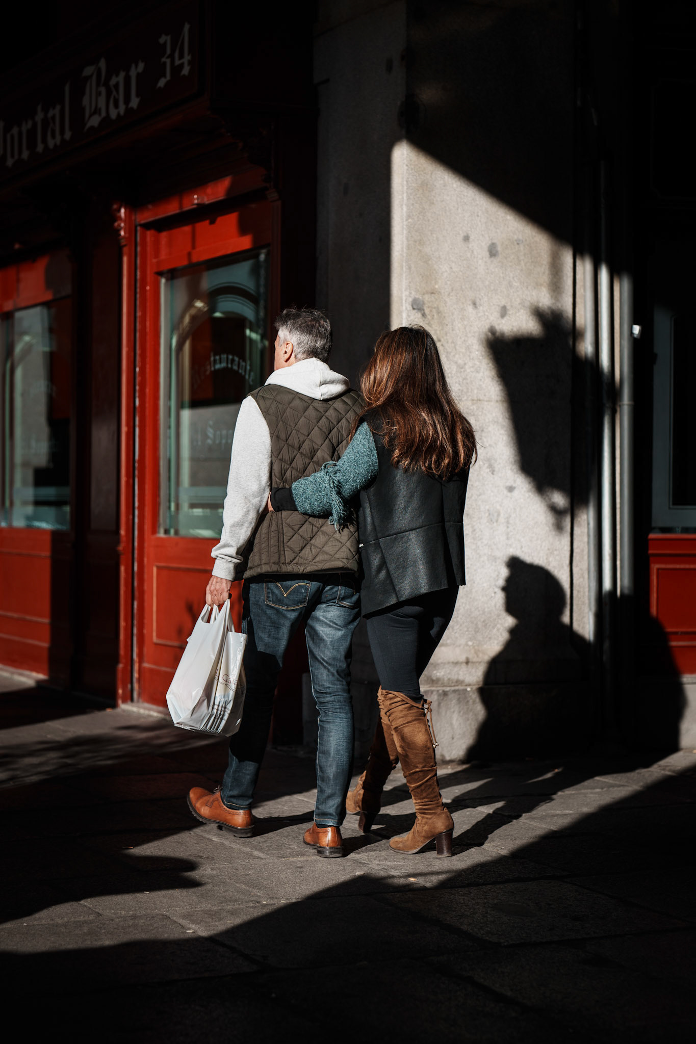 through the sunlight filtering under the arcades at plaza mayor, they walk together, close and quietly connected. the morning shadows stretch around them, creating shapes that tell their own story, a blend of light and form. her arm around his, his hand carrying the morning’s finds – small signs of a shared life and steady companionship. surrounded by the subtle warmth of madrid’s red façades and stone columns, they move in step, wrapped in their own rhythm amidst the hum of the city.