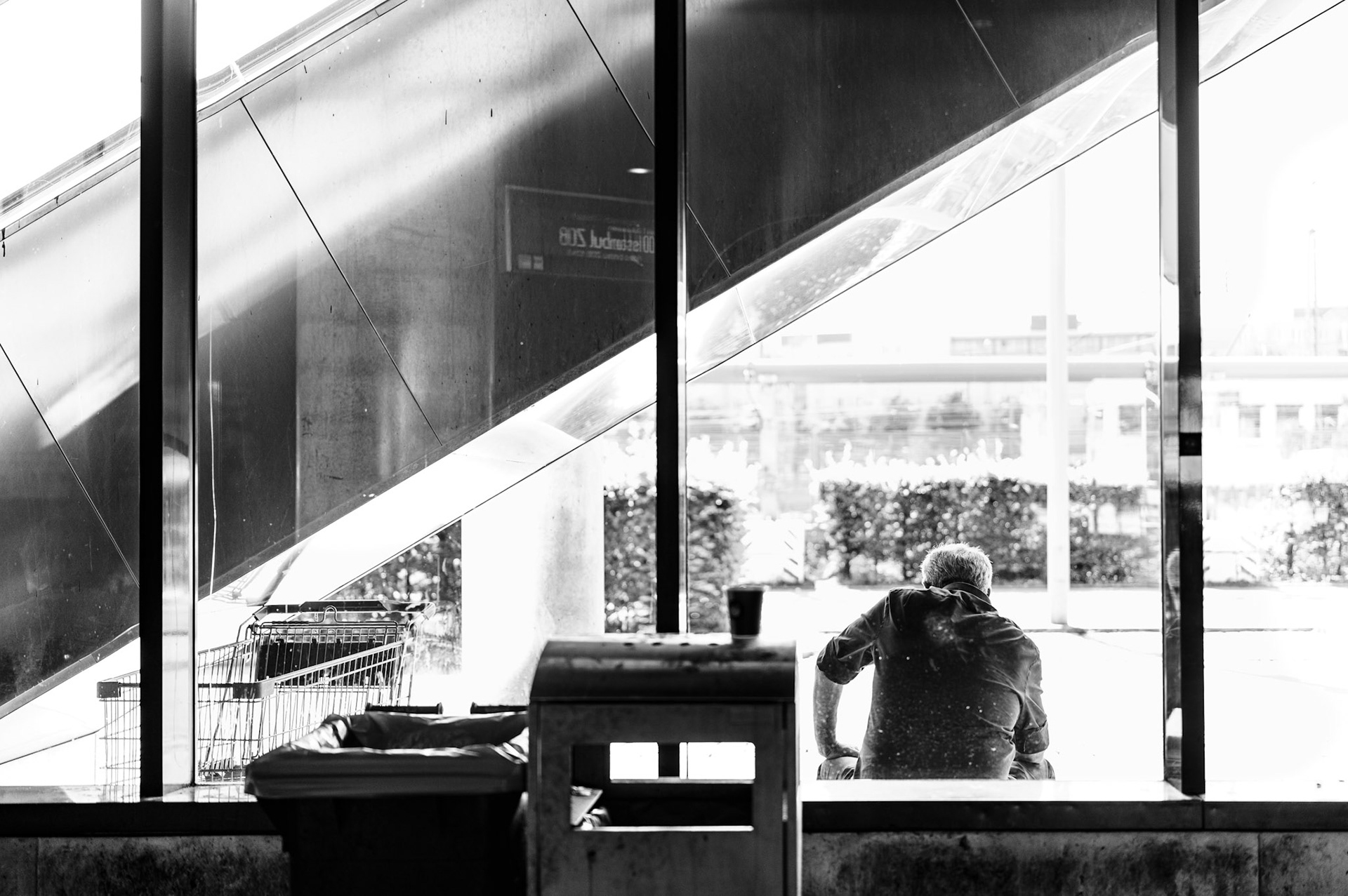 captured in a fleeting instant, this photo reveals a man sitting alone, framed by the geometric lines of a modern urban structure. the bright light outside contrasts starkly with the shadowy interior, creating a scene where the subject is almost silhouetted against the glass. the reflections, the textures of metal and glass, and the diagonal lines of the staircase add a graphic element that pulls the viewer’s eye through the frame. there’s a sense of quiet solitude, of a moment suspended in time—caught between the inside and the outside, the public and the private, the familiar and the unknown. it is an exploration of isolation within the busy, bustling cityscape.