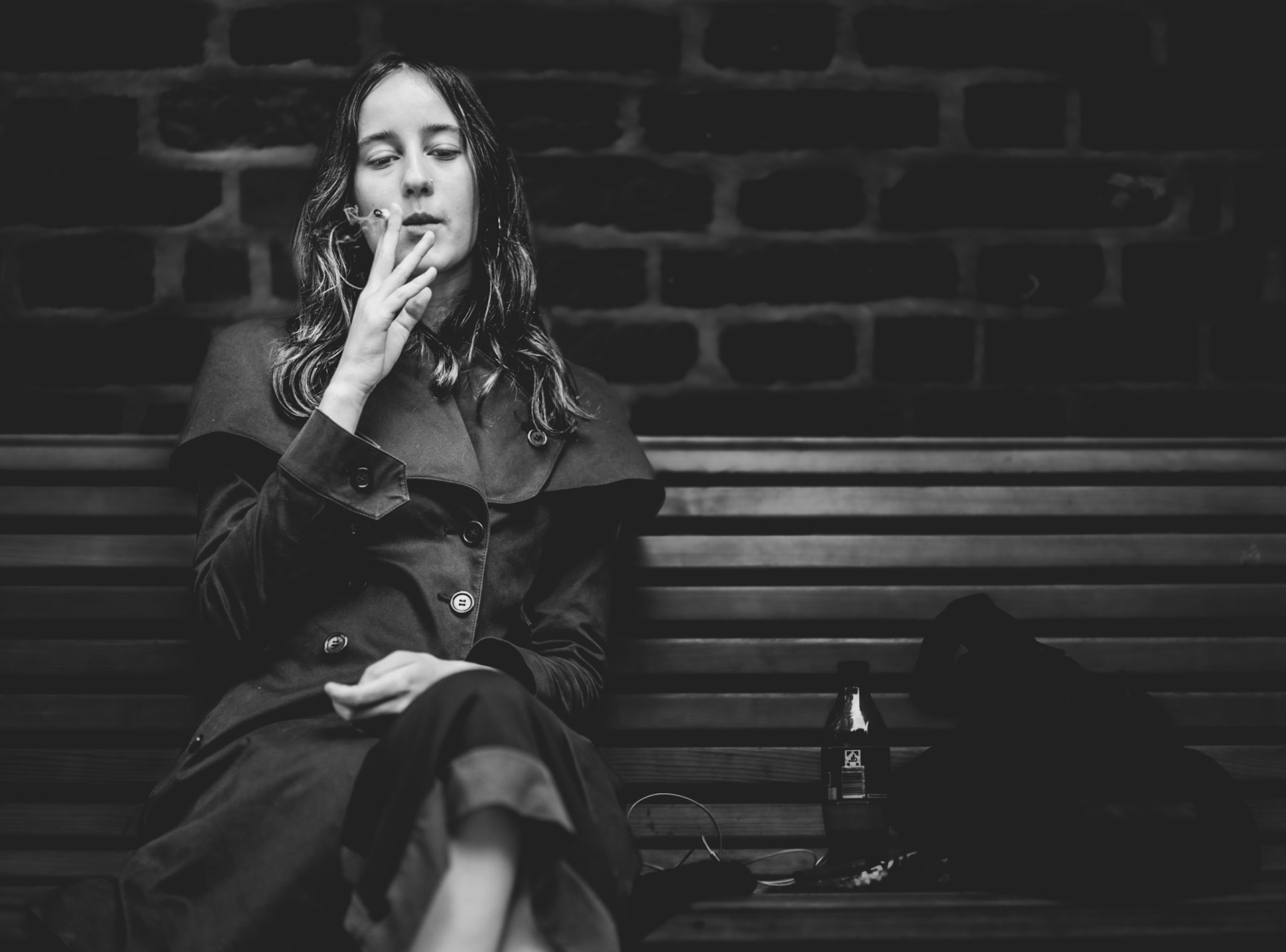captured in munich's city center, this photograph features a young woman sitting on a bench in front of an old brick church. her relaxed posture and contemplative expression, along with the cigarette in her hand, create a poignant scene of introspection and solitude. the black-and-white composition emphasizes the texture of the brick wall and the subtle details of her coat, highlighting the quiet intensity of the moment.