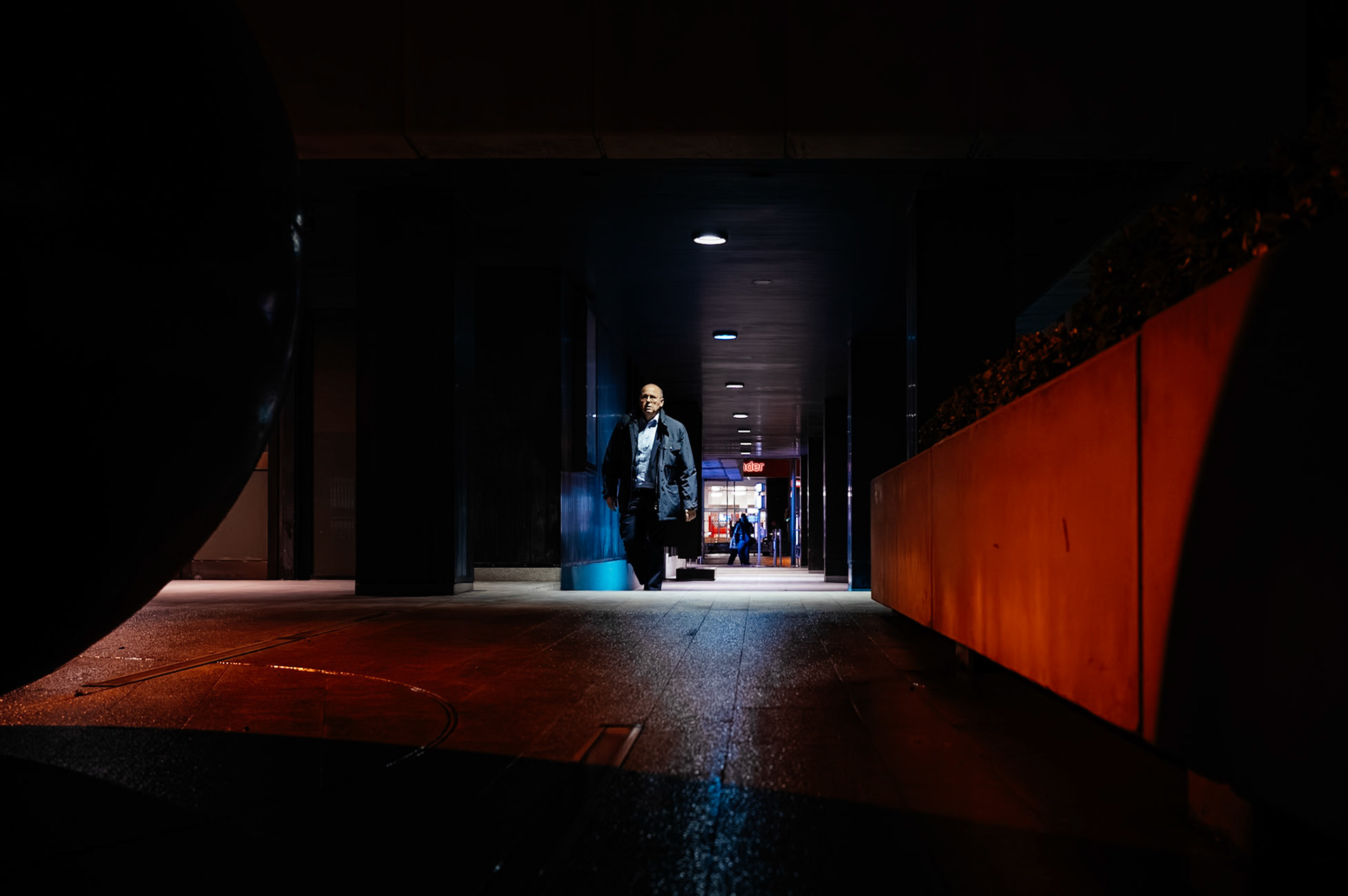 on a winter evening in the pere garau district of palma de mallorca, a lone man emerges from the shadows. bathed in the stark light of the urban landscape, he walks with purpose, his figure etched against the shop-lit corridor that leads into the heart of the city's night.