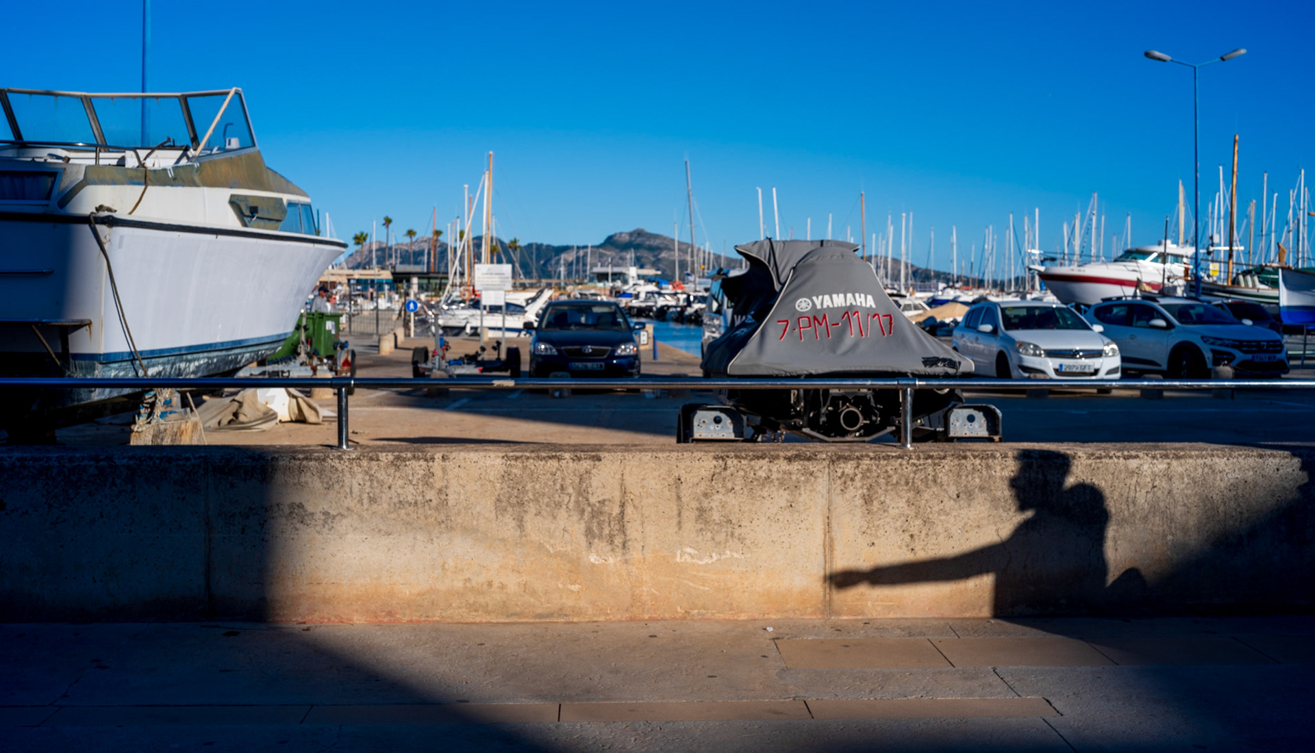 as the sun journeys across port de pollença's azure skies, it carves out a silent narrator on the wall: the shadow of a man, gesturing towards an unseen marvel. this shadow, a stark silhouette against the warm concrete, tells a story of engagement and wonder, as if the man is pointing out a chapter in the port's own tale. his outstretched arm could be directing our gaze to the sailboats nodding in the breeze, to the distant peaks, or to the simple yet profound beauty of life by the sea. every person and object becomes an actor on the stage of the marina, under the artful direction of light and shadow.