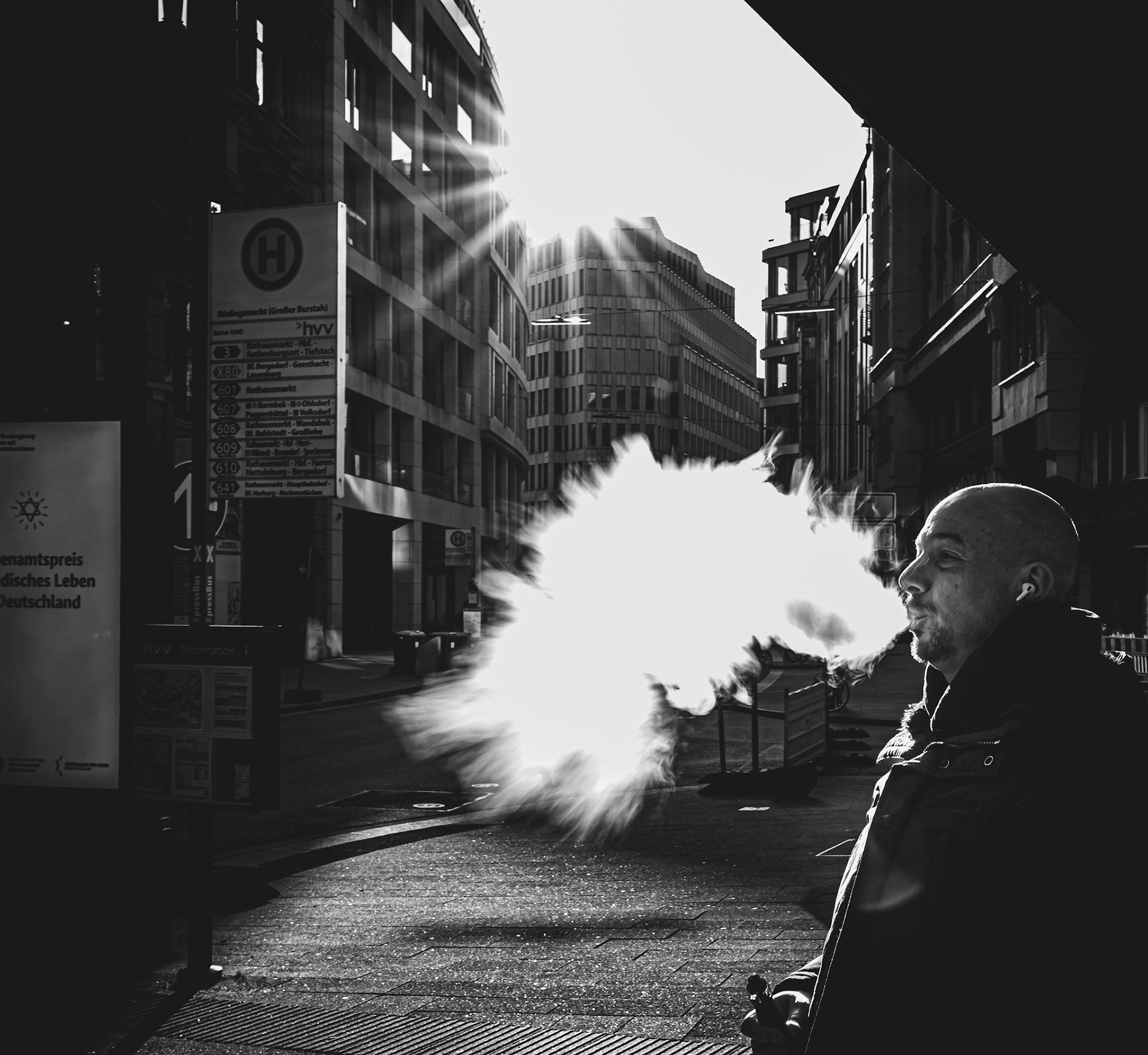 in the brisk hamburg dawn, a man stands enveloped in sunlight, a cloud of smoke escaping into the cold air. as the city stirs, this solitary figure captures a moment of calm before the day's rhythm escalates. the rising sun's rays carve light through the urban landscape, heralding the new day.
