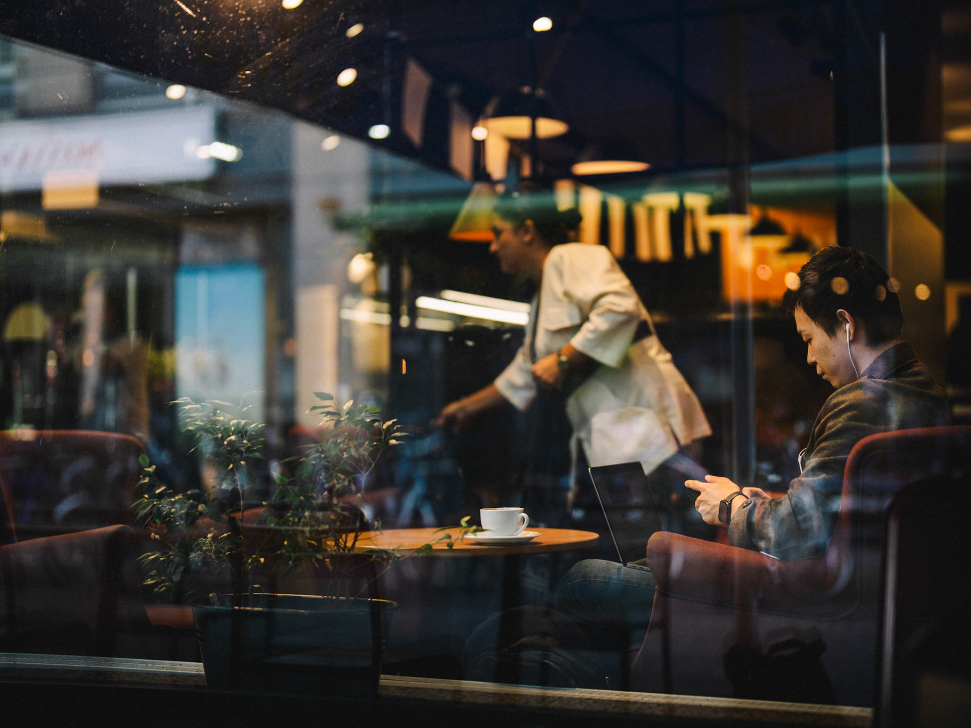 captured through the café window in berlin mitte, this image reveals a young man immersed in his own thoughts, separated from the bustling world outside. the warm reflections and soft colors of the café atmosphere evoke a sense of calm and introspection, as if he’s momentarily detached from his surroundings. the glass creates a quiet barrier between observer and observed, between outside and inside, while the light gently highlights the contours of his face and hand. this moment of silent reflection tells a small, personal story amidst the big city, as if the world around him has paused for just a second.