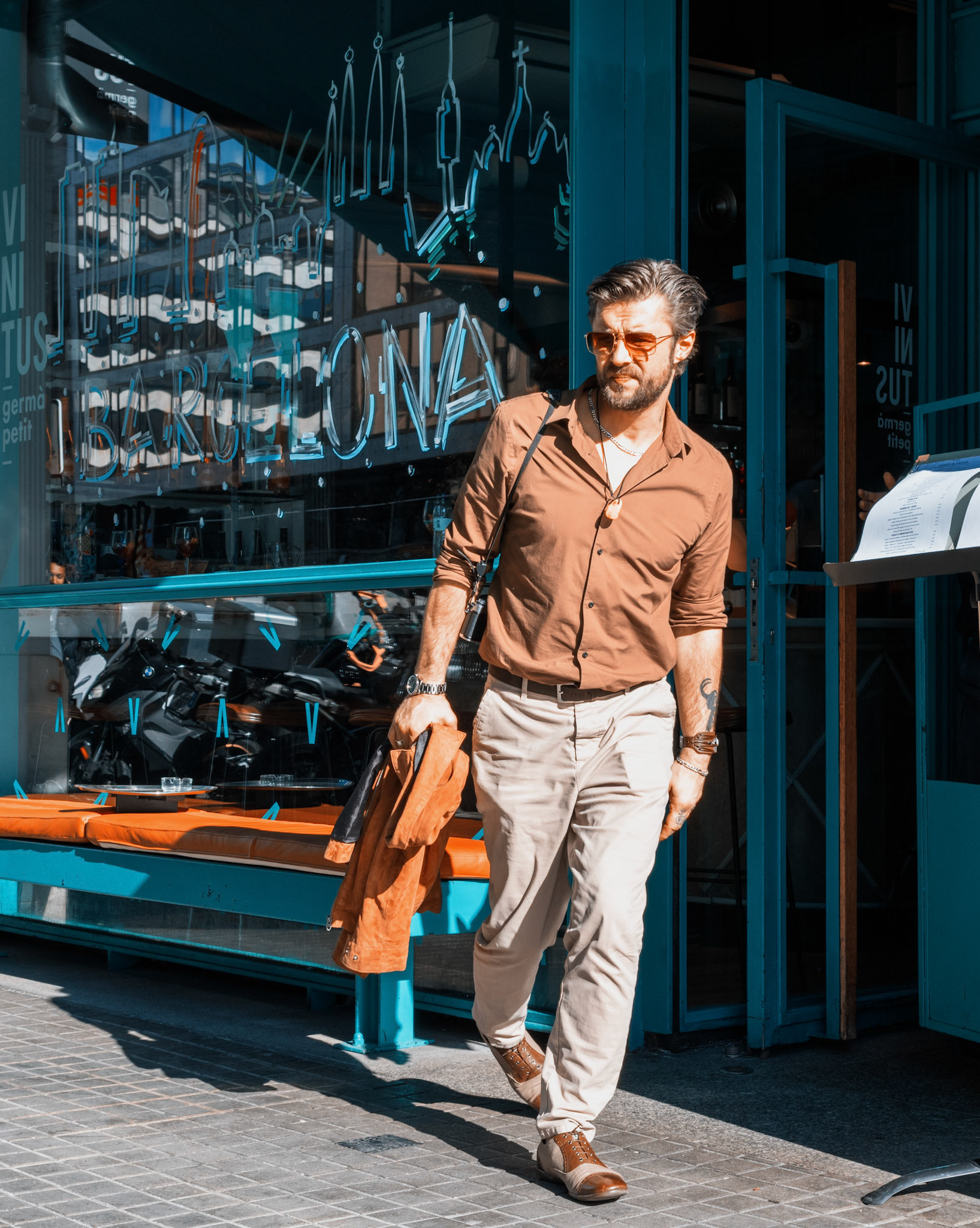 captured in barcelona, where the vibe of the city pulses through the streets, a man exits the resstaurant vinitus with a stride reflecting the casual elegance of the catalan capital. his style speaks without words, a narrative of confidence woven into the fabric of his shirt, the drape of his jacket. shades rest on his face, not just to shield from the mediterranean sun, but as an accent of his persona. the blue hues of the doorway behind him seem to announce his presence, a quiet proclamation of sophistication. as he walks, the tattoos peek from under his sleeves, hints of stories etched in ink, adding layers to the man's enigmatic allure. every step is a brushstroke in the portrait of a city that dresses not just in fashion, but in an attitude of composed assurance.