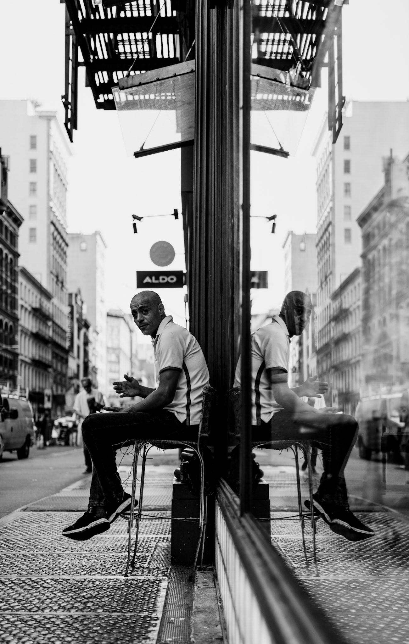 captured on broadway, this photograph features a man taking a brief respite. just as he notices the camera, he acknowledges it with a smile, embodying a moment of spontaneous connection and perhaps a lesson in openness for us all. the reflection in the window creates a captivating symmetry, emphasizing the urban environment and the human element within it. this candid shot highlights the beauty of everyday encounters in the city.