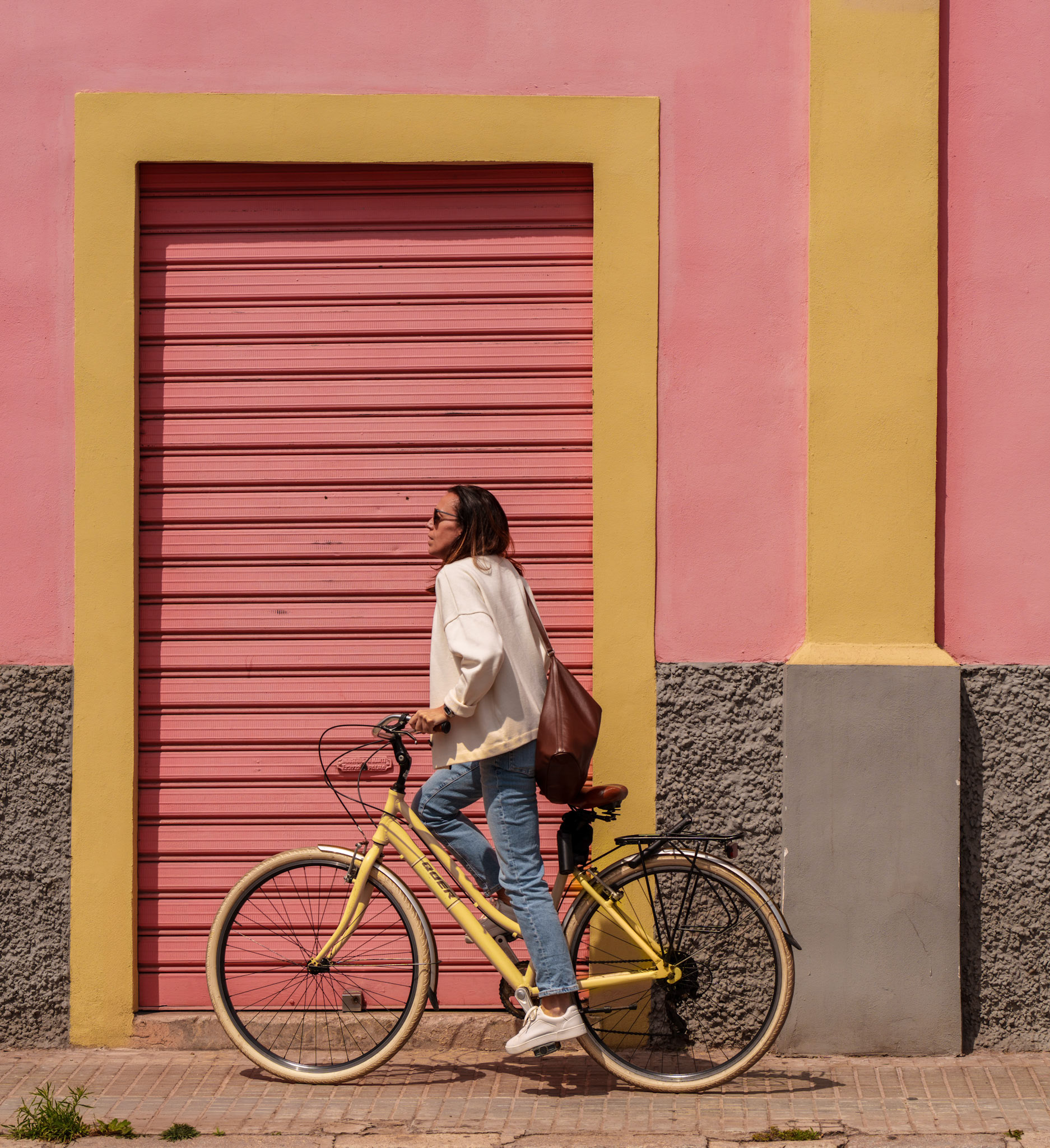 in the vibrant district of el terreno, where the quaint charm of colorful yet untouched houses whispers stories of old, she lingers. here, against a backdrop of candyfloss walls, a moment rests, suspended in the warmth of the sun. the peach blush of the rolling shutter, the mellow yellow framing it, and the coral hue of the facade, they come together like a painter's deliberate stroke on a canvas craving life. the bicycle, a pastel echo to the scene, bears a rider dressed in the softest of whites, her silhouette a serene contrast to the geometry around her. she's poised as if caught between destinations, a still life within the city's gentle heartbeat.