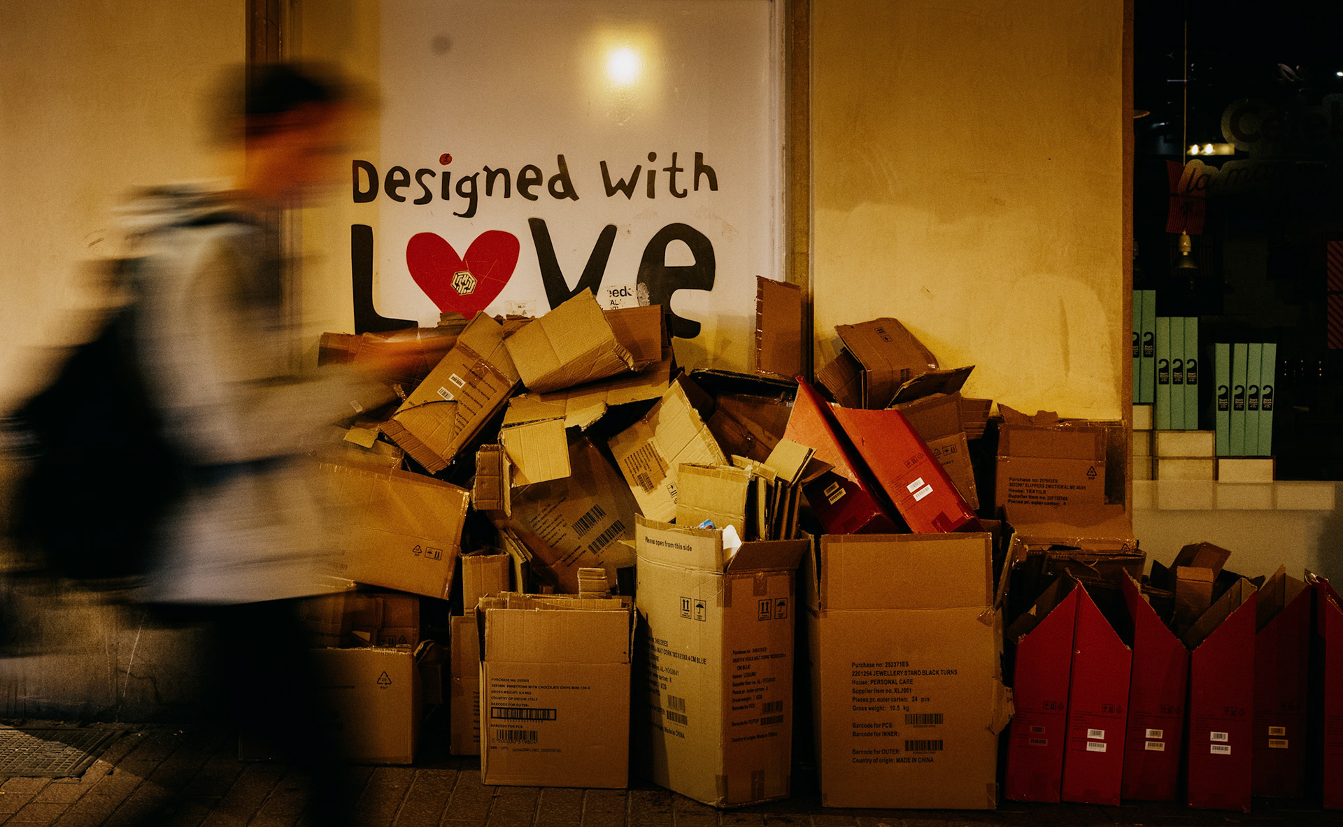 night folds its velvet curtain around the narrow streets of palma’s old town, where golden hues spill from the storefronts, casting a warm glow on a scene less ordinary. a passerby, a ghostly blur of motion, dances by a mountain of cardboard boxes piled up like an urban still life. it’s as if each box, with its own story, now rests in the after-hours of consumption, stamped with the irony of a window sign that exclaims “designed with love.” there's poetry in this juxtaposition, a silent commentary on the transient treasures we hold dear by day, discarded by night. the photo feels like a still from a film, one that captures the essence of life’s fleeting nature, a moment of transition frozen in time. the city breathes, its heartbeat palpable in the quiet shuffle of feet against cobblestone, the murmur of distant chatter, the gentle rustle of paper in the wind.