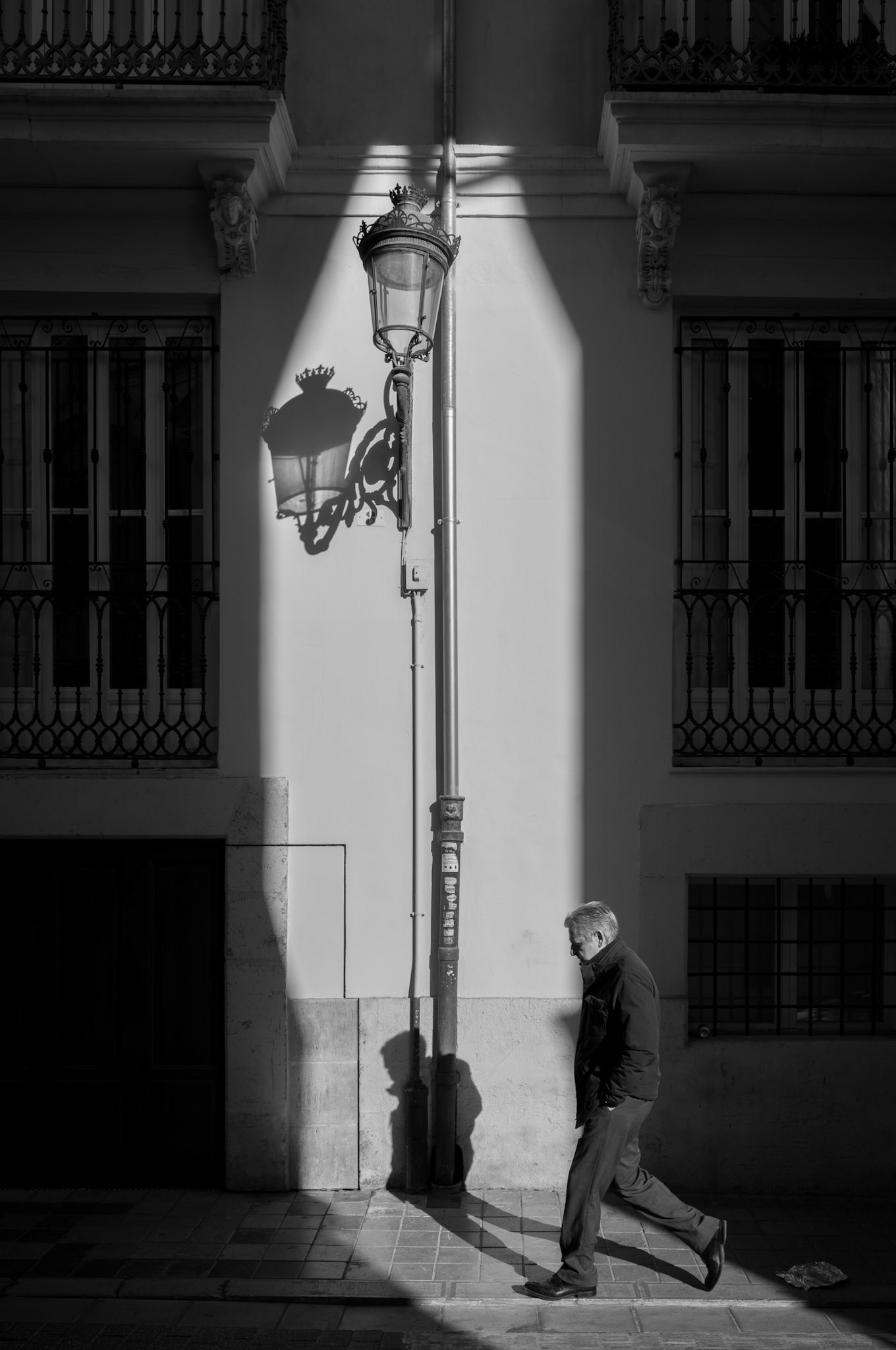 as the sun peeks through a slender gap between the age-old edifices of valencia, it orchestrates the day's first spectacle. a street lamp, still lit as a lingering sentinel of the night, is caught in a stark burst of dawn. its shadow, a crisp, dark echo on the sun-washed wall, tells of the city's awakening. the man, emerging from the architecture's embrace, walks alongside his own elongated silhouette, a fleeting companion in the dance of light. this moment, a brief intersection of past and daybreak, invites contemplation of the day's nascent potential.