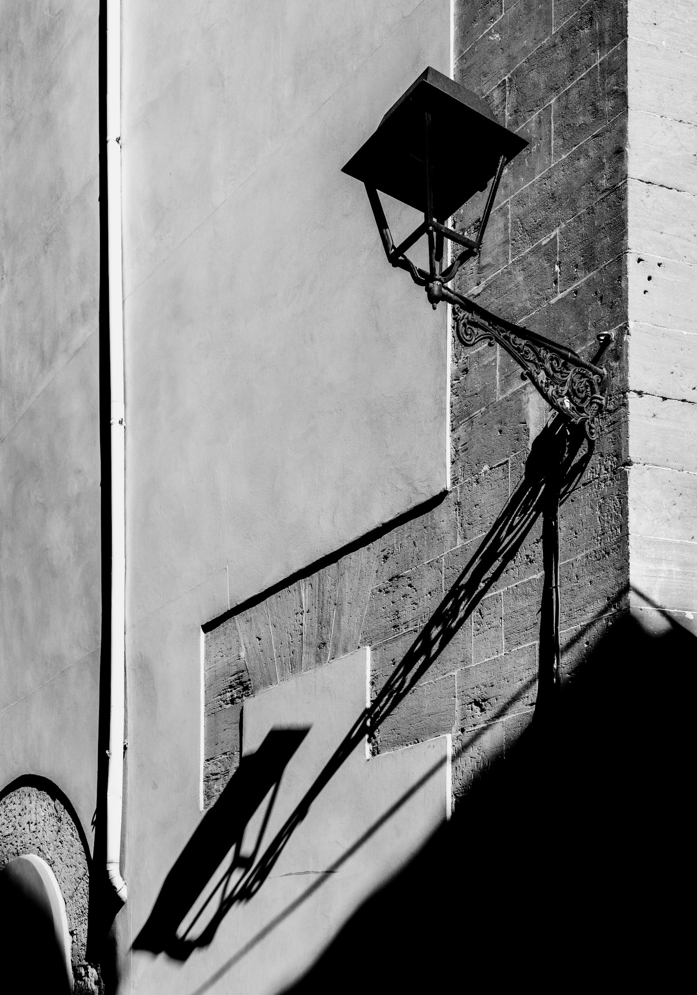 in the historic district of la lonja, where the whispers of the sea breeze through narrow alleys, i captured this interplay of light and shadow. the old street lamp, with its ornate ironwork, casts a sharp silhouette against the textured stone wall. this area, though now bustling with tourists, retains its romantic charm with scenes like this—simple yet evocative, merging the past with the present. the stark contrast and clean lines in this black and white photograph evoke a sense of timelessness, highlighting the architectural beauty and serene ambiance of palma's beloved neighborhood.