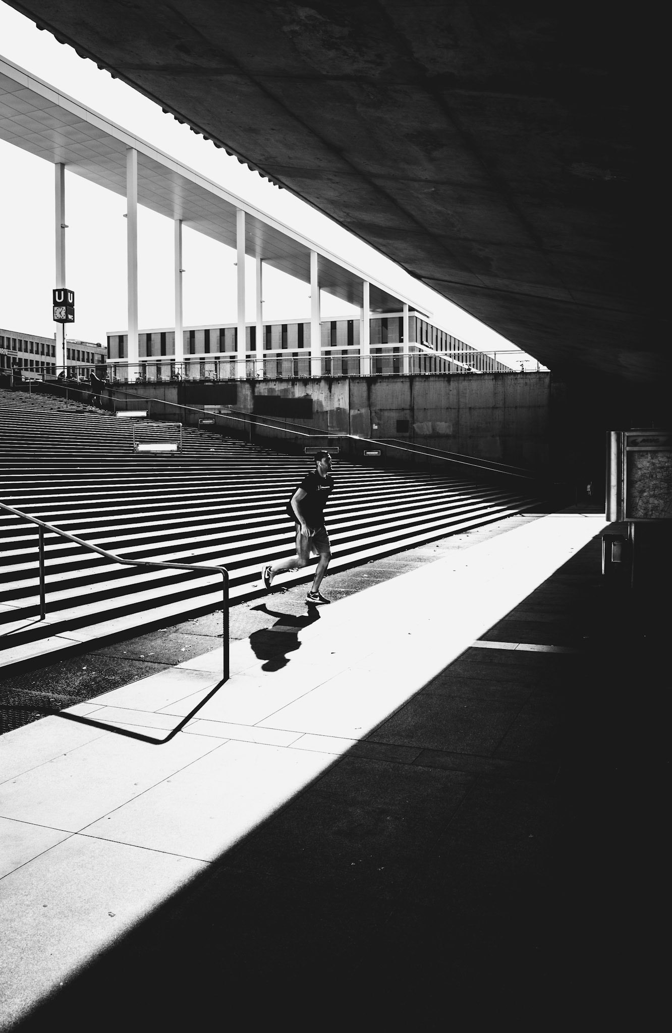 on a bright afternoon, the cityscape turns into a stage, and he runs through the spotlight, chased by his own shadow. the architecture frames his movements, and the shadows cast create a rhythm that captures the fleeting dance of light. beneath the concrete and steel, a human figure in motion, a reminder of life in the starkness of urban geometry.